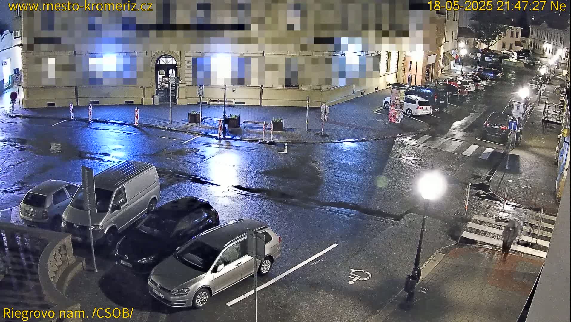A wet, nighttime street scene shows several parked cars, a van, and a few pedestrians near a building.