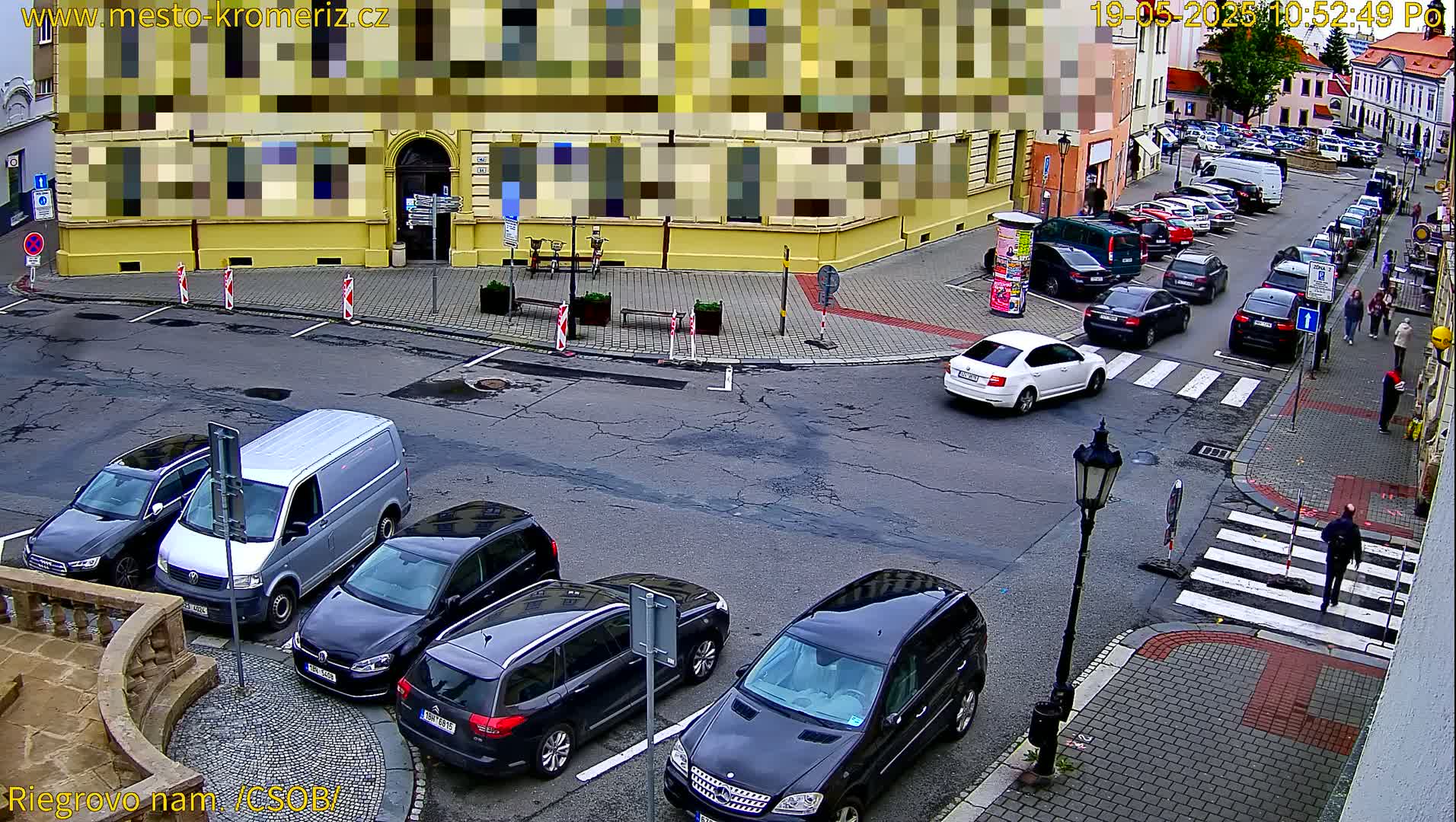 A street corner view shows several parked cars and a few cars driving on a street with a pedestrian crossing, under an overcast sky.