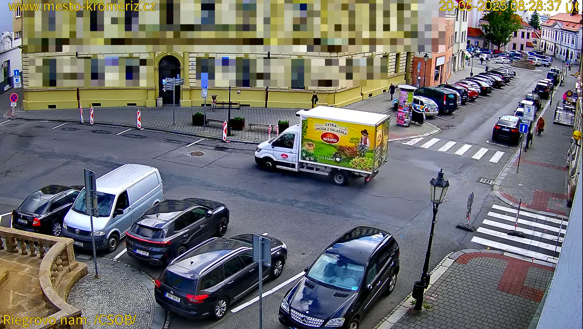 A street scene on a sunny day shows several cars parked along the curb and a few more in a parking lot, near a white delivery truck with advertisements, and a pedestrian crossing.