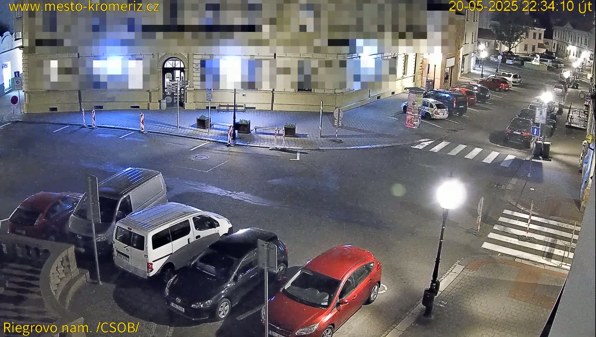 At night, several cars are parked in a lot next to a building, with a crosswalk visible in the foreground under streetlights.