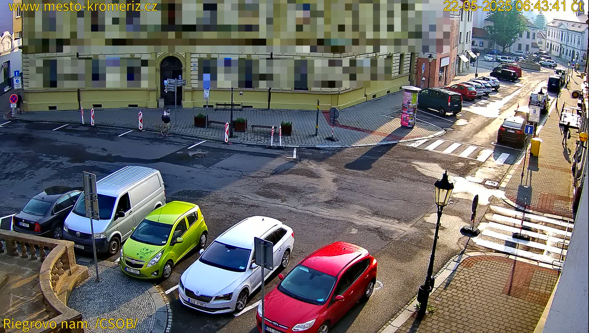 A sunny street corner shows several parked cars, a van, and pedestrians near a building.