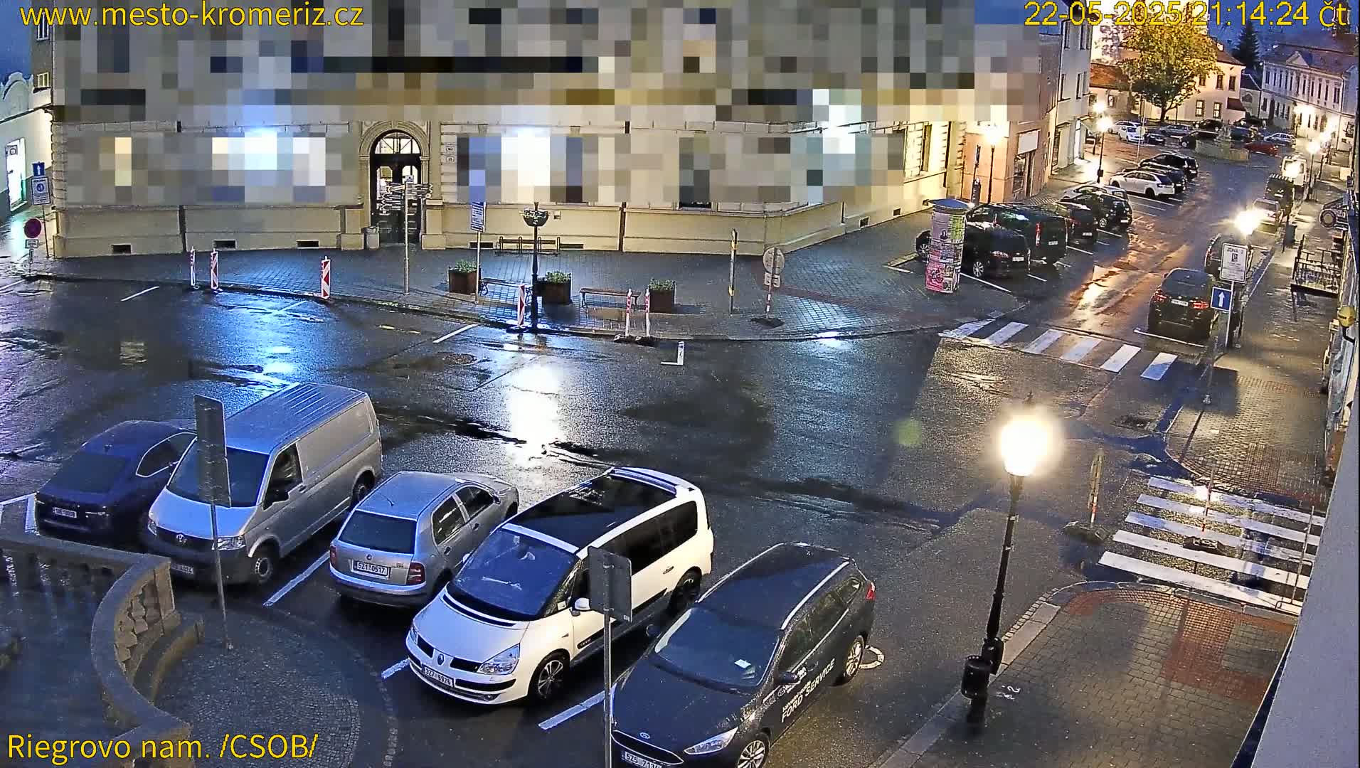 A wet, nighttime street scene shows several cars parked in a town square, with some cars driving through the intersection.