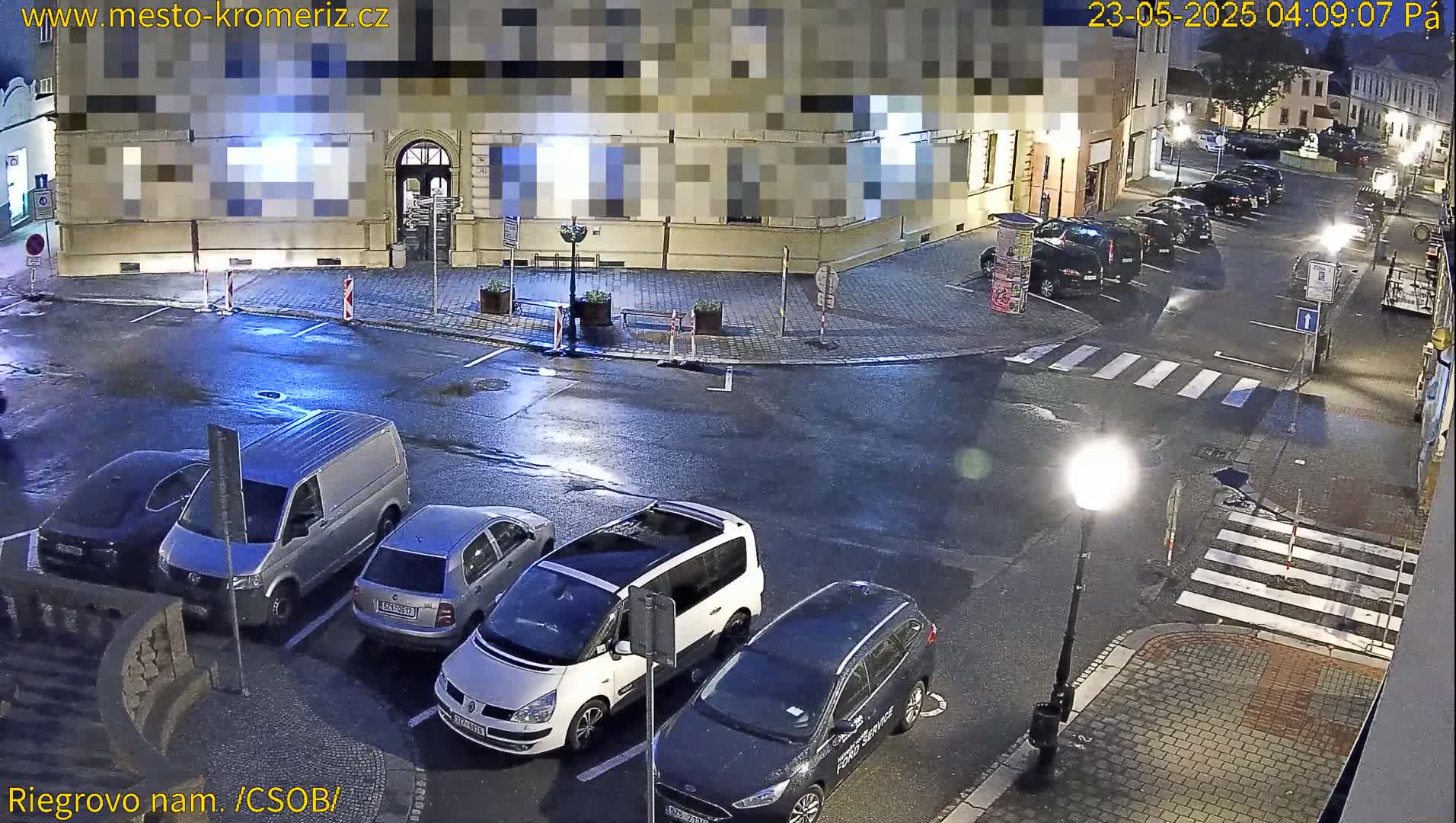 A wet, nighttime street scene shows several cars parked in a lot next to a building, with a pedestrian crosswalk visible in the foreground.