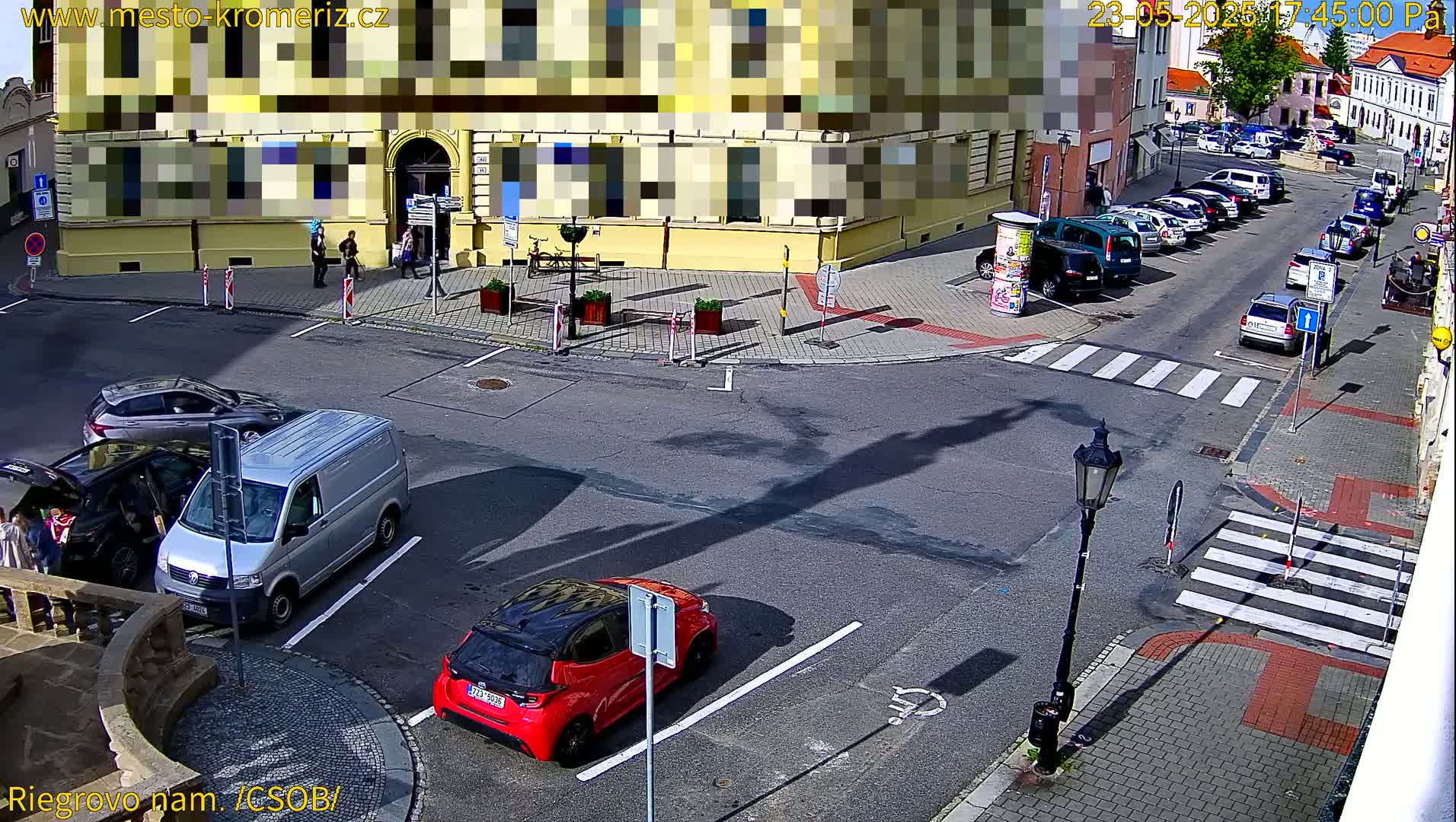 A sunny street scene shows several cars parked and driving around a corner near a yellow building, with a few pedestrians nearby.
