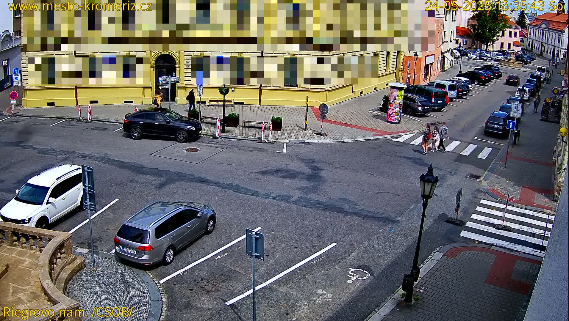 A sunny day in a town square shows several cars parked and driving around a corner, with pedestrians crossing a crosswalk.