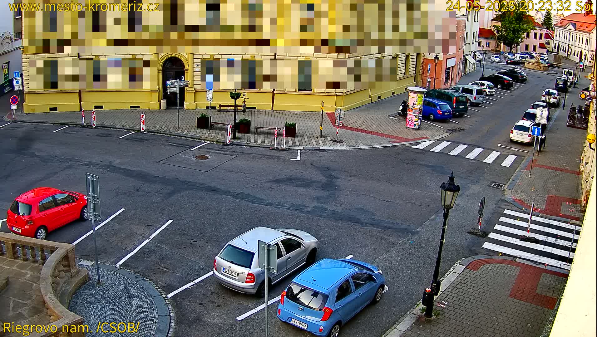 A street corner view shows several parked cars, a pedestrian crossing, and a yellow building with partially obscured signage; the weather appears clear.