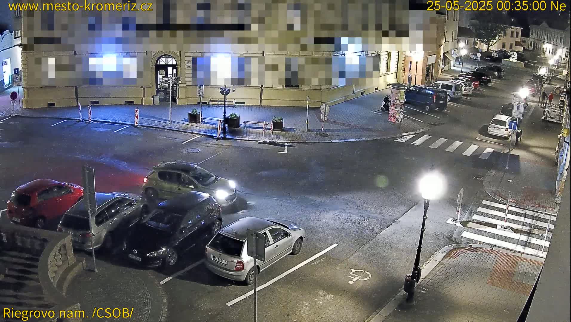 At night, several cars are parked in a lot near a building and crosswalk under streetlights.