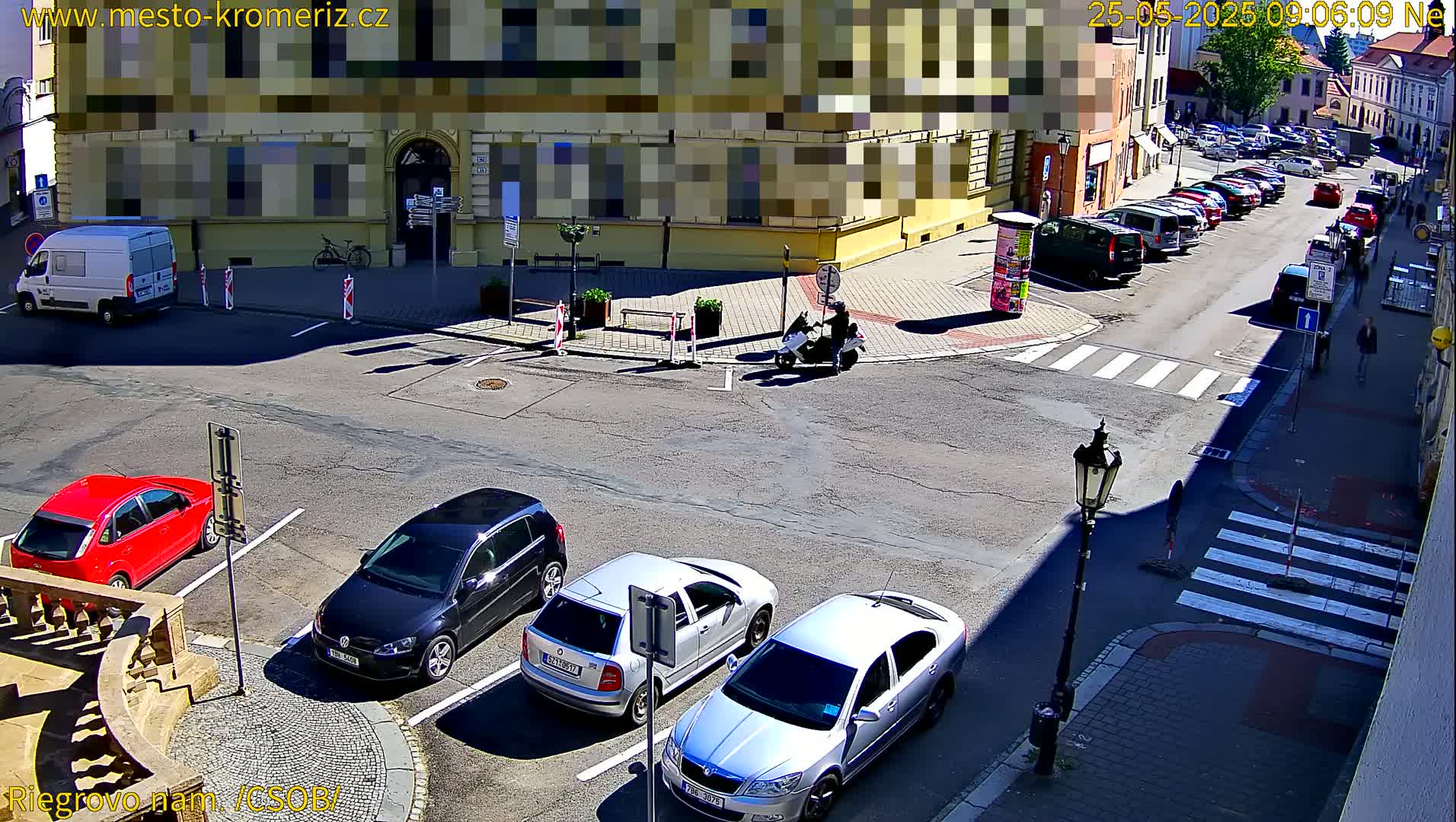A sunny street scene shows several cars parked in a lot and along a curb, with a scooter and a van also visible, near a building and a crosswalk.