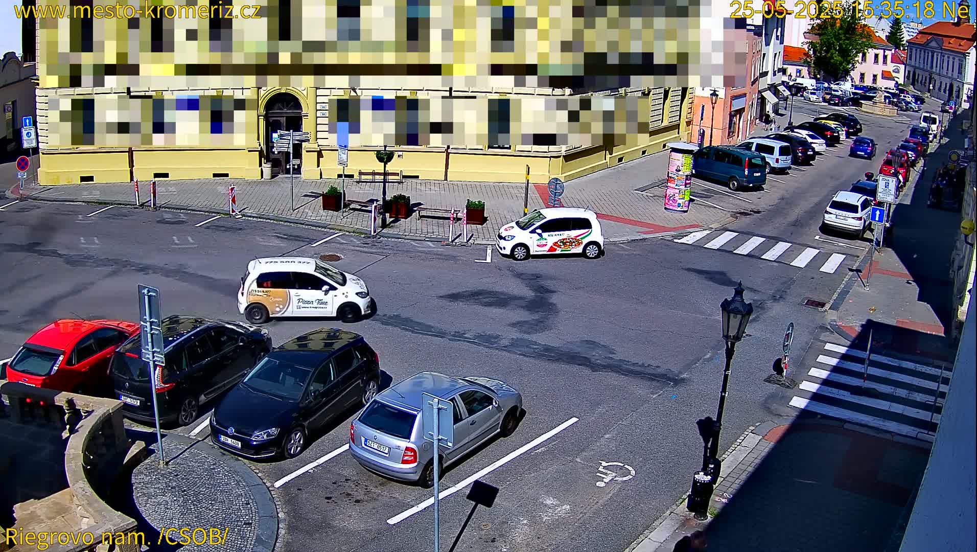 A sunny day reveals a town square with several cars parked in a parking lot and others driving on the street, near a yellow building.