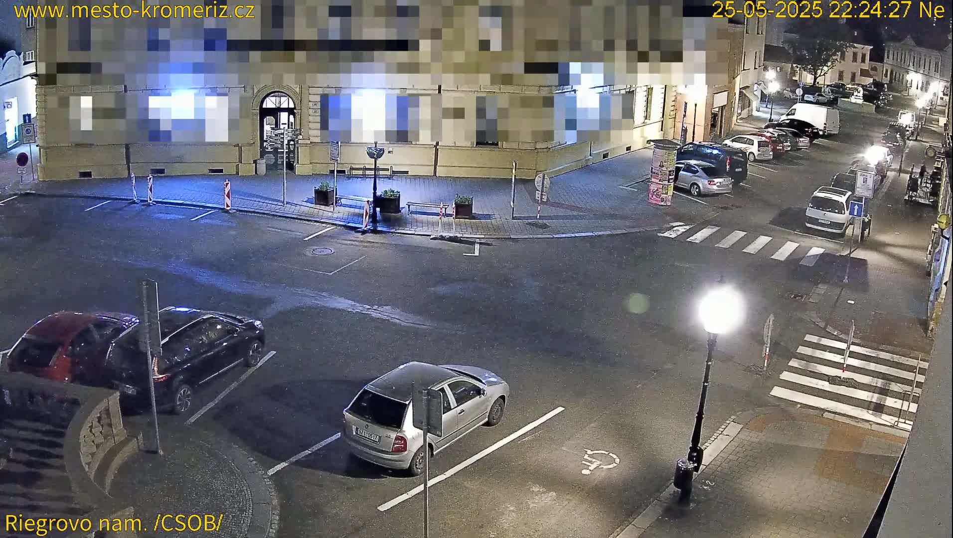 A nighttime, high-angle view of a town square with several parked cars, a building, and a pedestrian crossing.