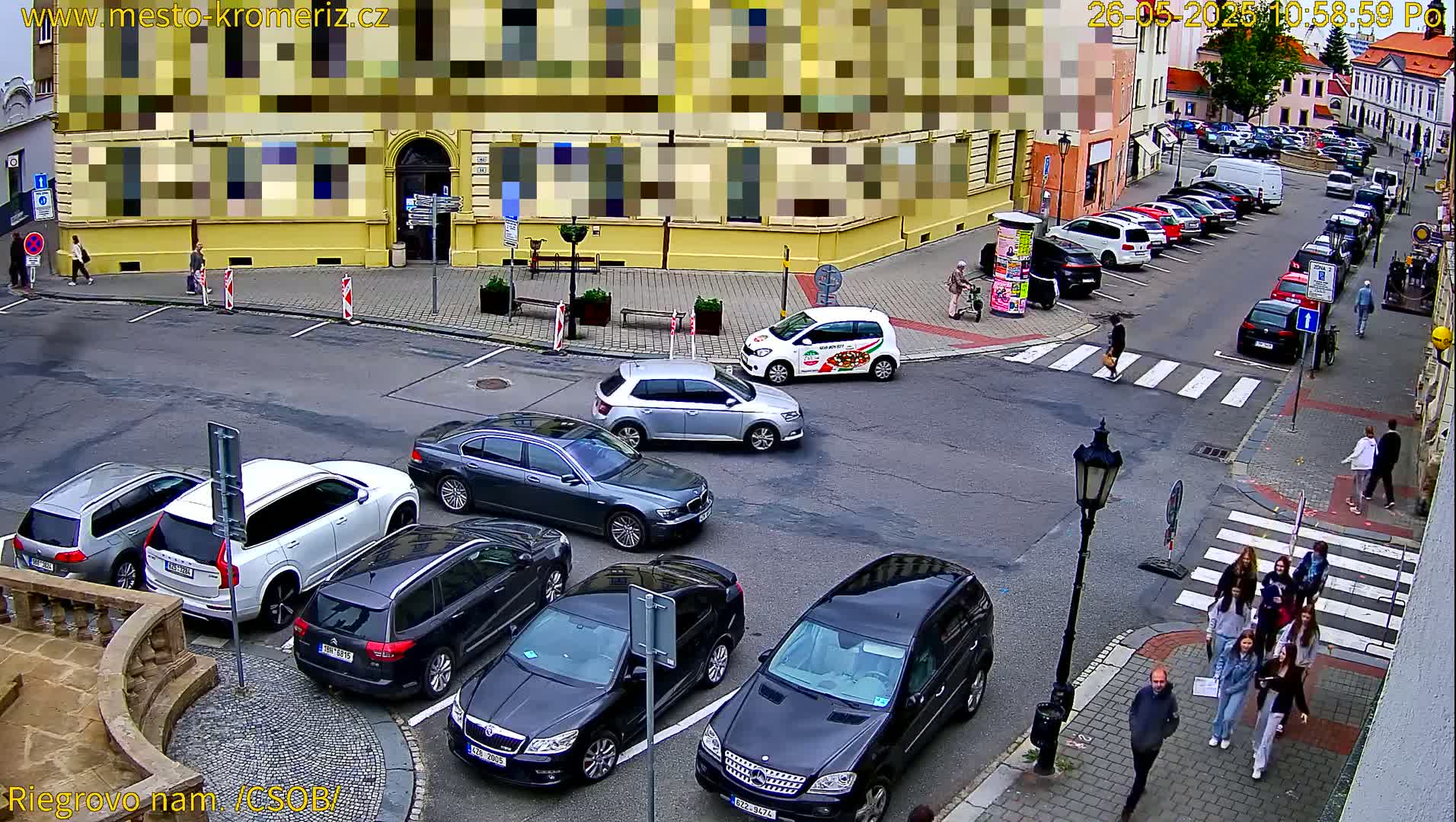 A street scene with parked cars and pedestrians, on a partly cloudy day.