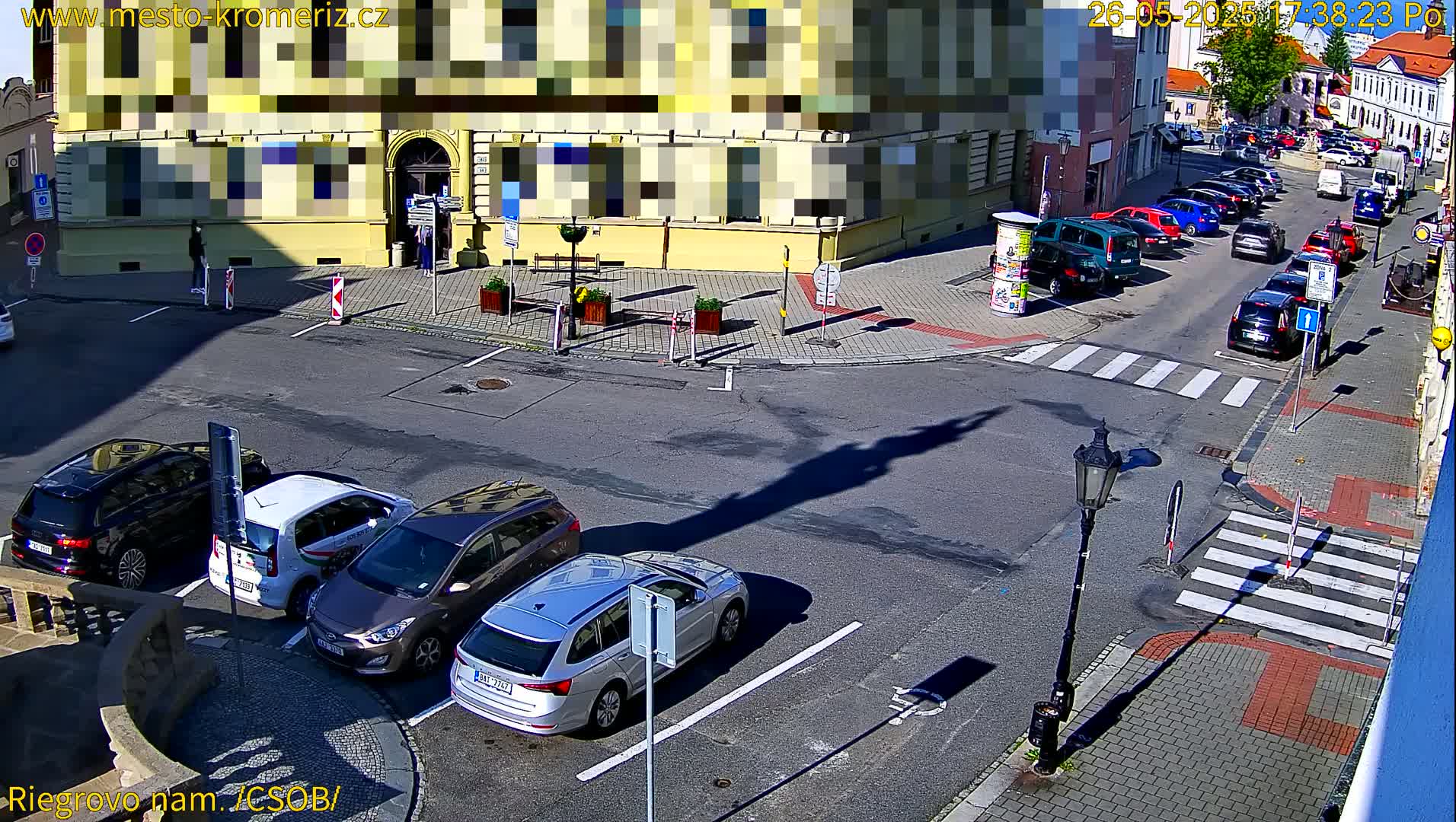 A sunny day view of a town square with several cars parked in a lot and more cars driving on the street,  along with a building and a lamppost casting shadows.