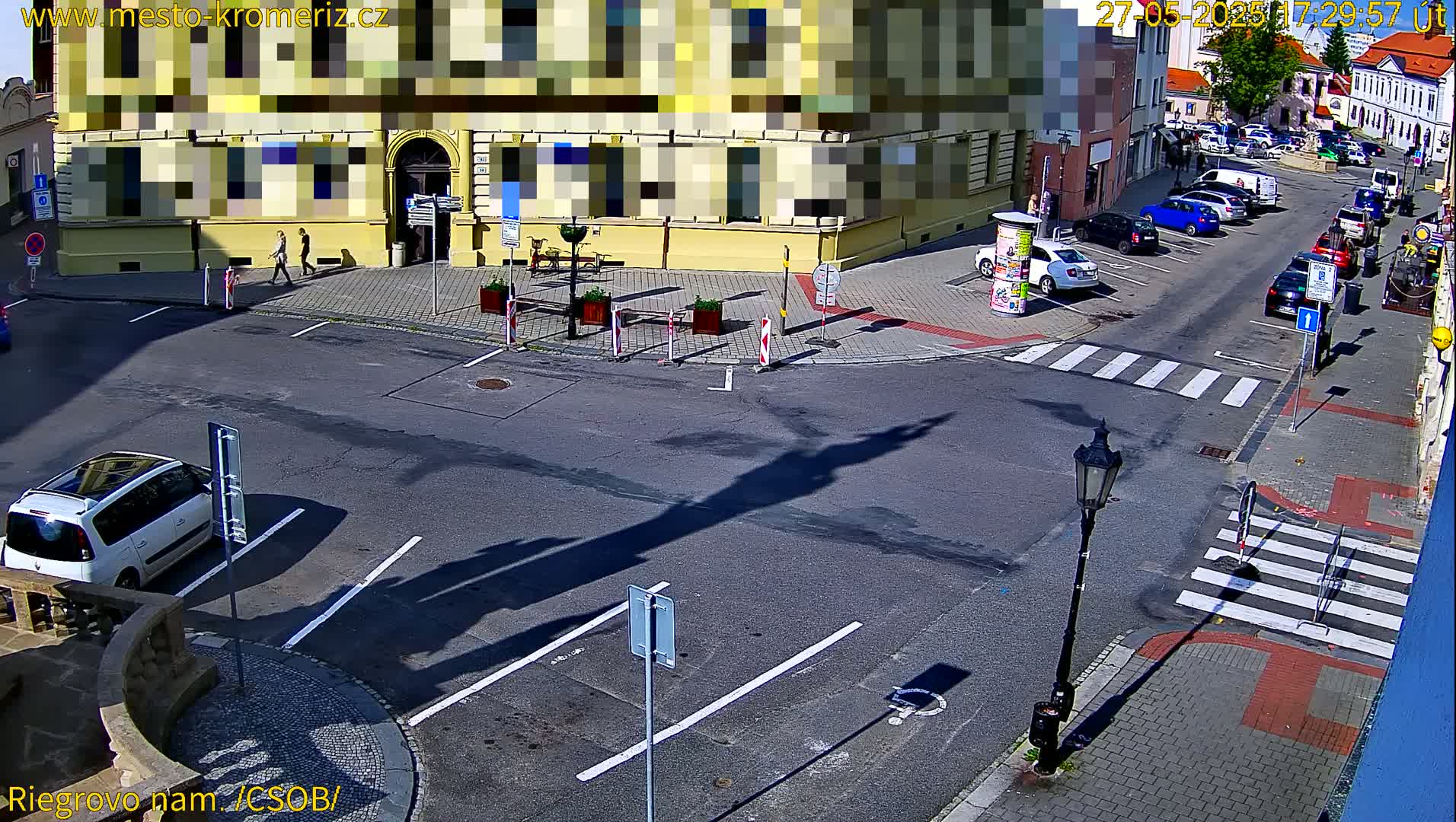A sunny street scene shows a mostly empty parking lot, a crosswalk, and several cars parked along the side of the road near buildings.