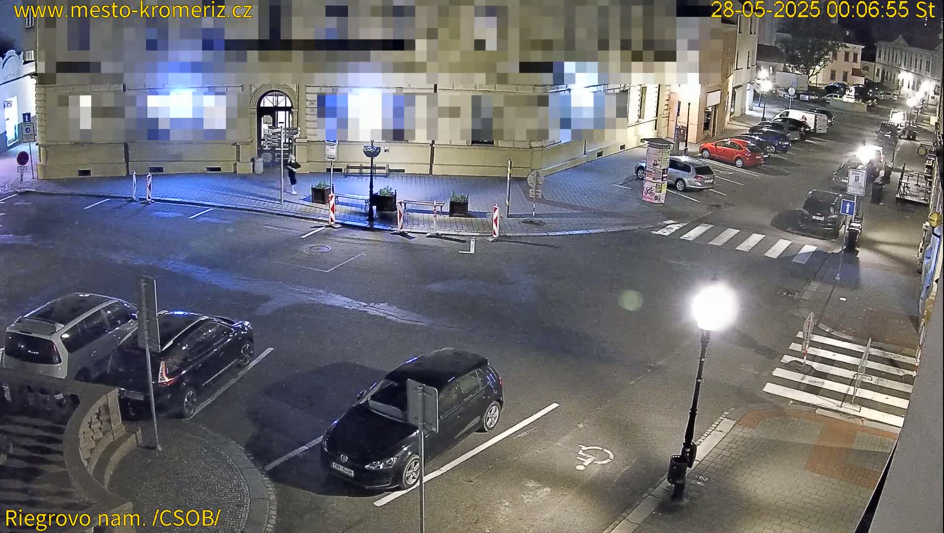 At night, several cars are parked in a lot near a street corner with a crosswalk under a streetlight.
