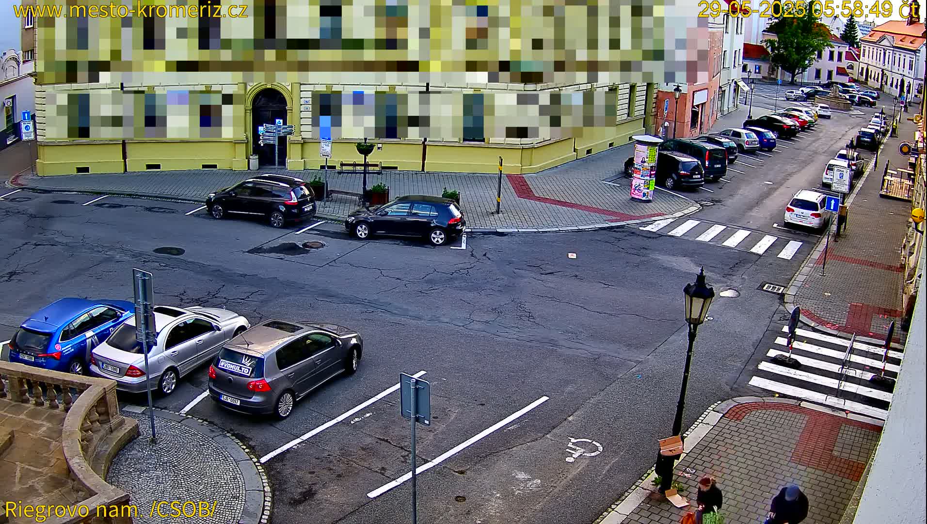 A street-level view of a town square with several parked cars and pedestrians, on a partly sunny day.
