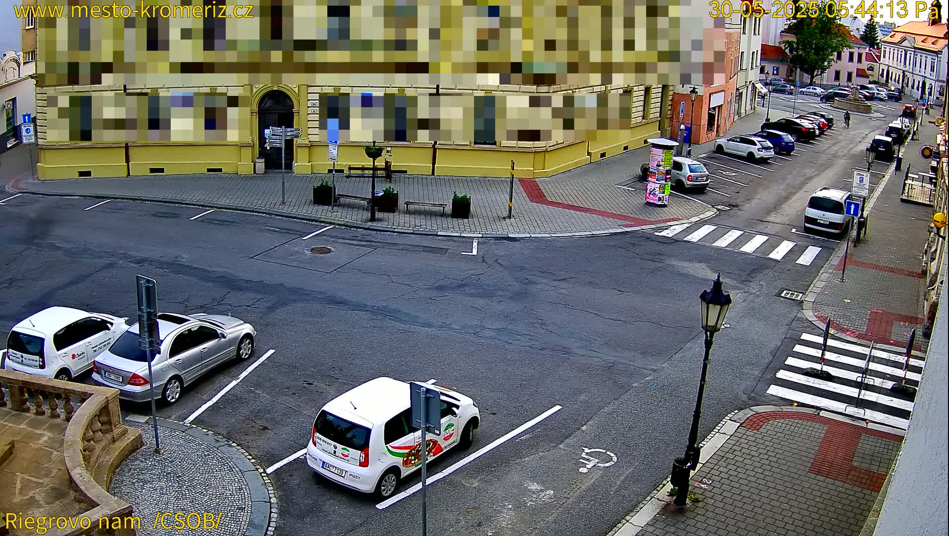 A street corner with parked cars and a few pedestrians is viewed from above on a partly cloudy day.