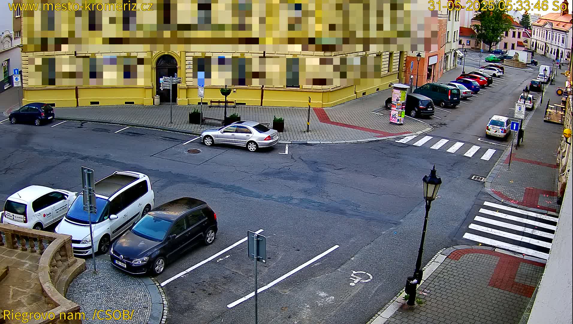A street corner with several parked cars and a few cars driving by, on a partly sunny day.