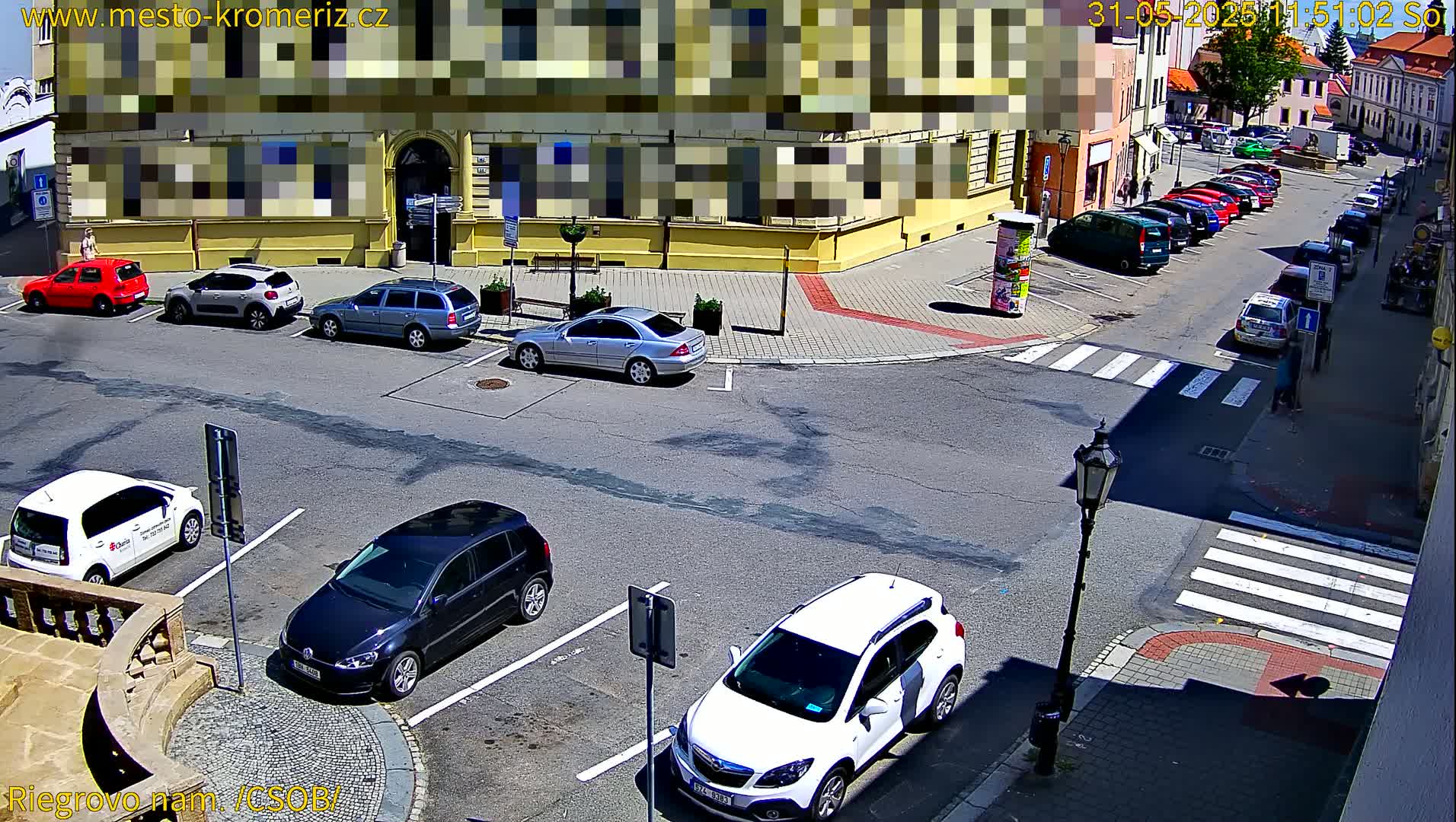 Several cars are parked in a sunny city square near a building, with a pedestrian crossing in the foreground.