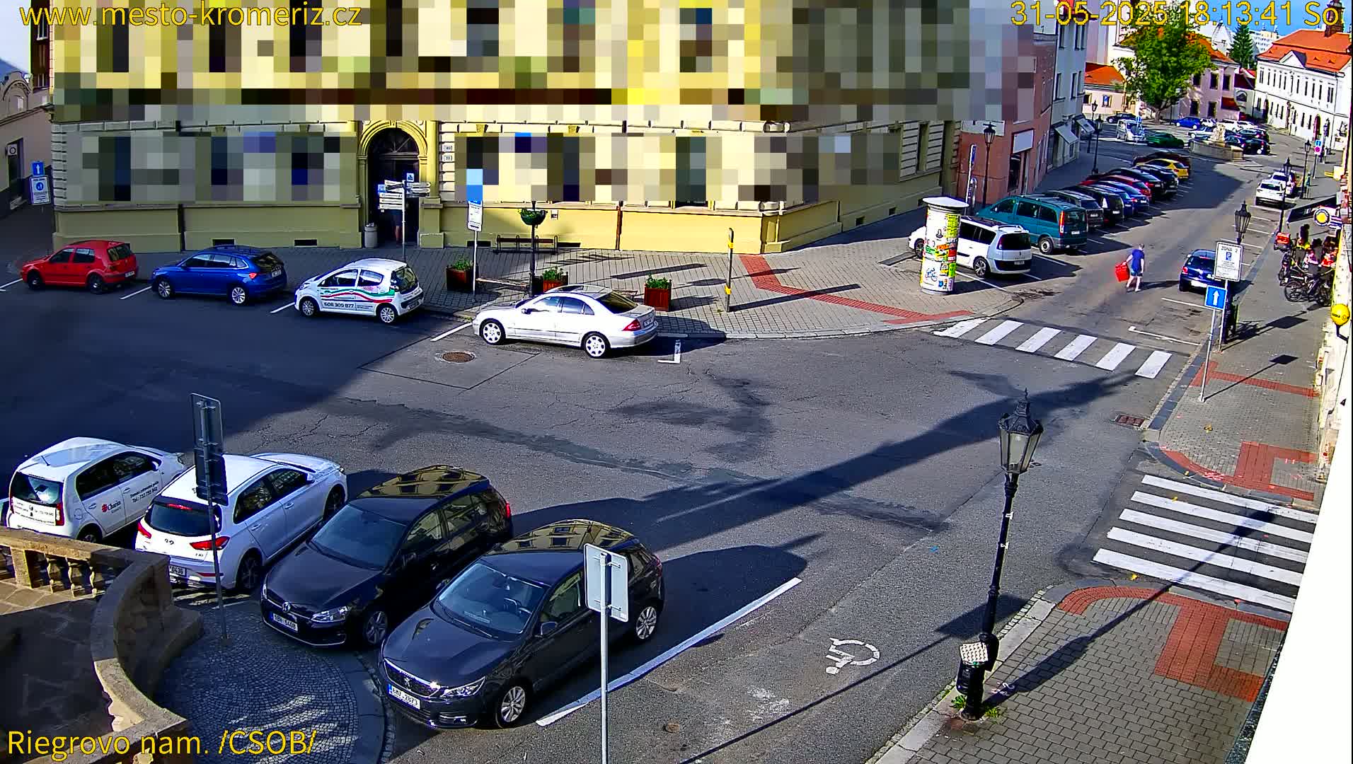 Several cars are parked on a sunny street corner near a building, with additional cars parked along a street in the background.