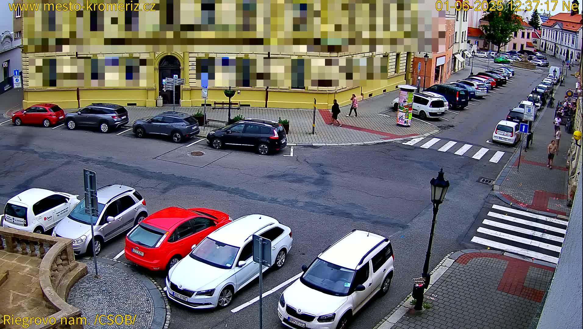 A street-level view of a parking lot and street intersection with several cars parked, a few pedestrians walking, and a lamppost, on a sunny day.