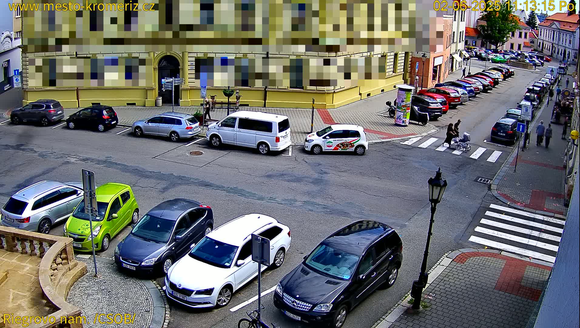 A street scene on a sunny day shows several cars parked along the curb and several others parked in a lot; pedestrians are crossing a crosswalk.
