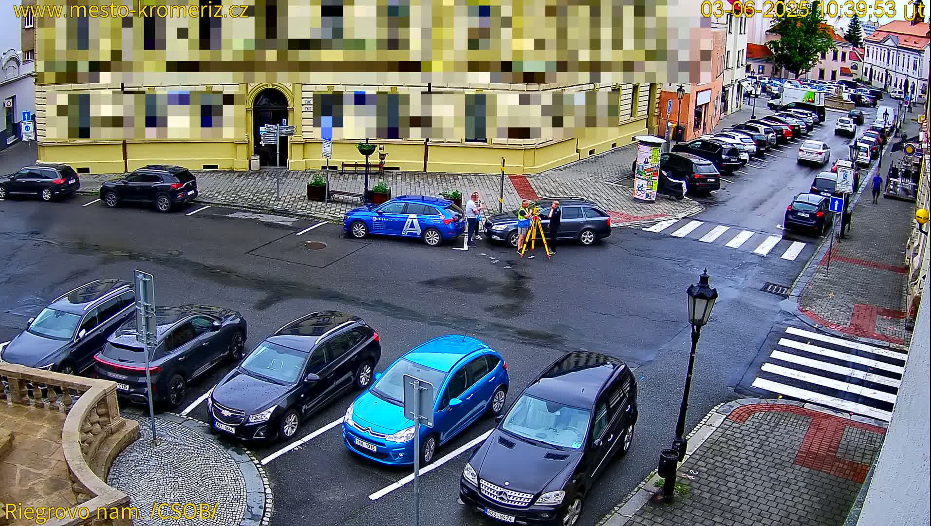 A wet city street scene shows several parked cars surrounding a small group of people near a surveying instrument, with additional cars parked along a street in the background.