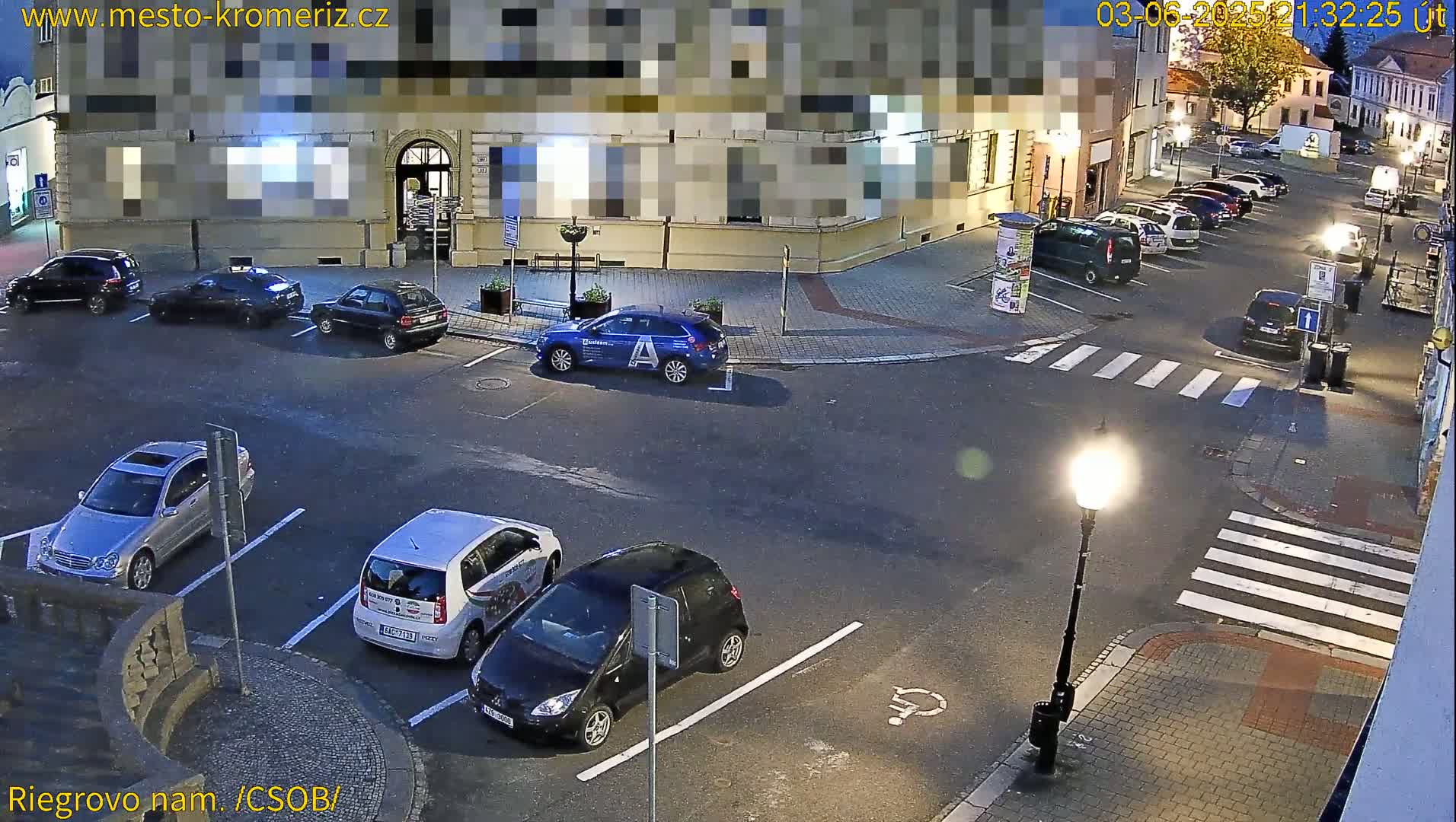 A nighttime, street-level view shows several cars parked in a lot and driving on a street next to a building under dim streetlights.