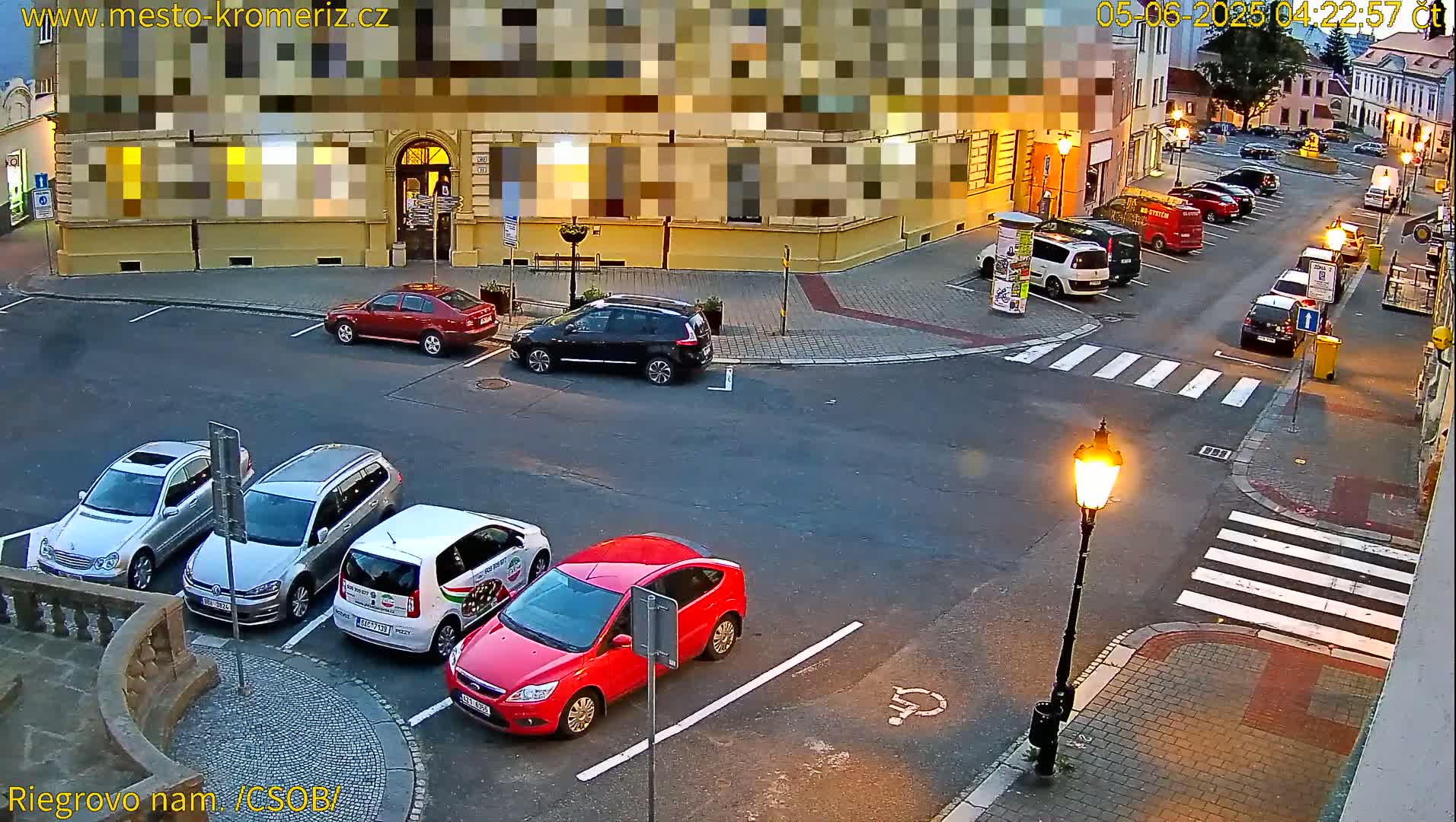 A town square at dusk, with several cars parked in a lot and along the street, under a mostly clear sky.
