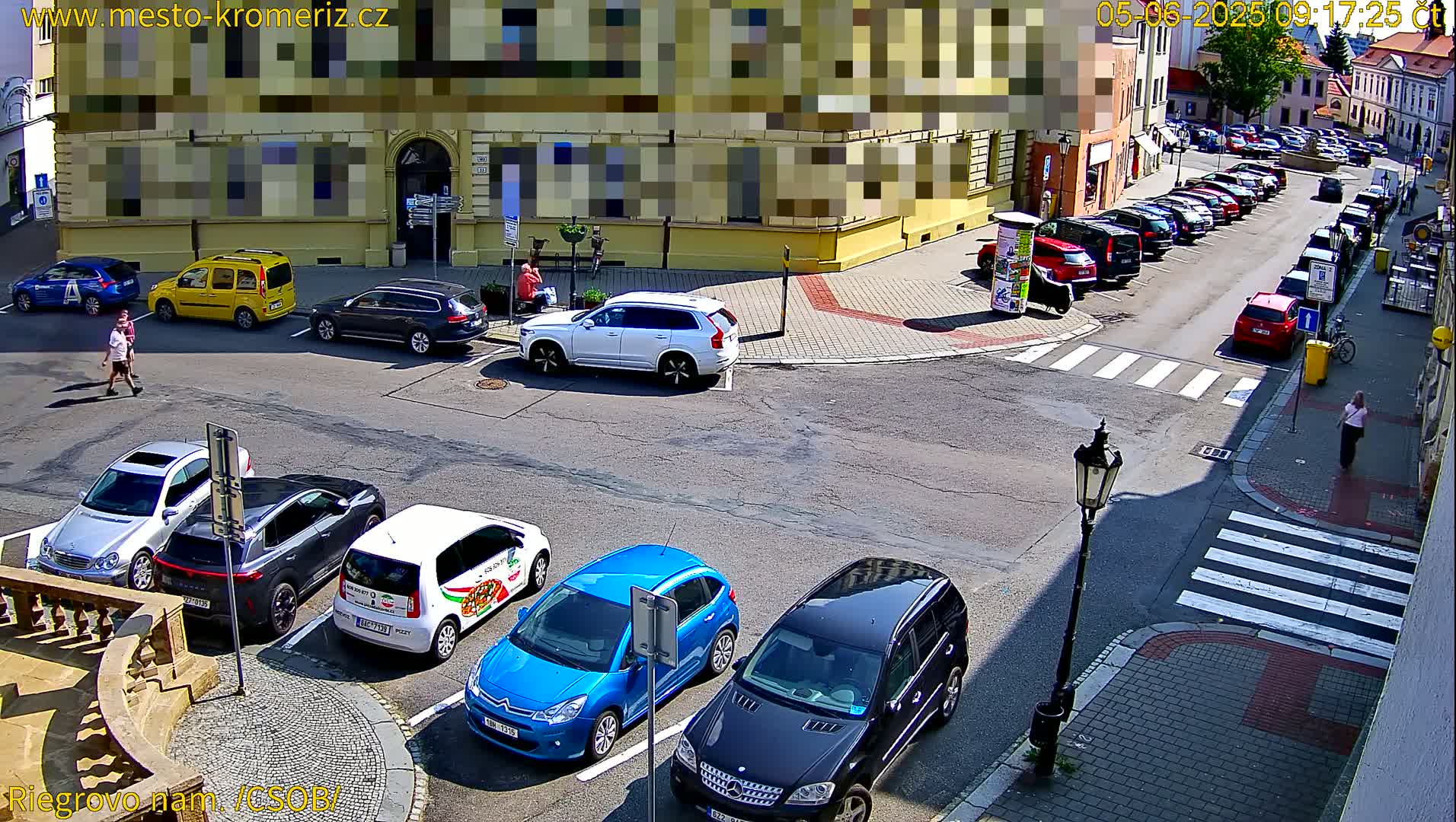 A sunny street corner shows several parked cars and pedestrians, with additional cars parked along the side of a street in the background.