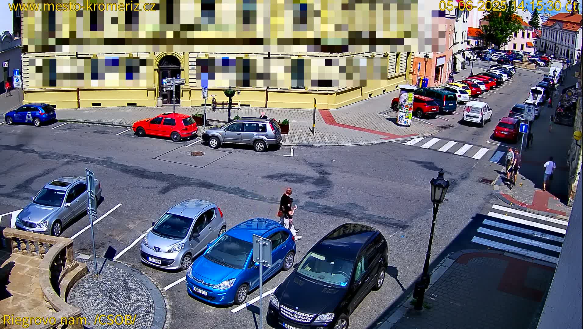 A sunny street scene shows a parking lot with several cars, a crosswalk with pedestrians, and a row of parked cars along a building.