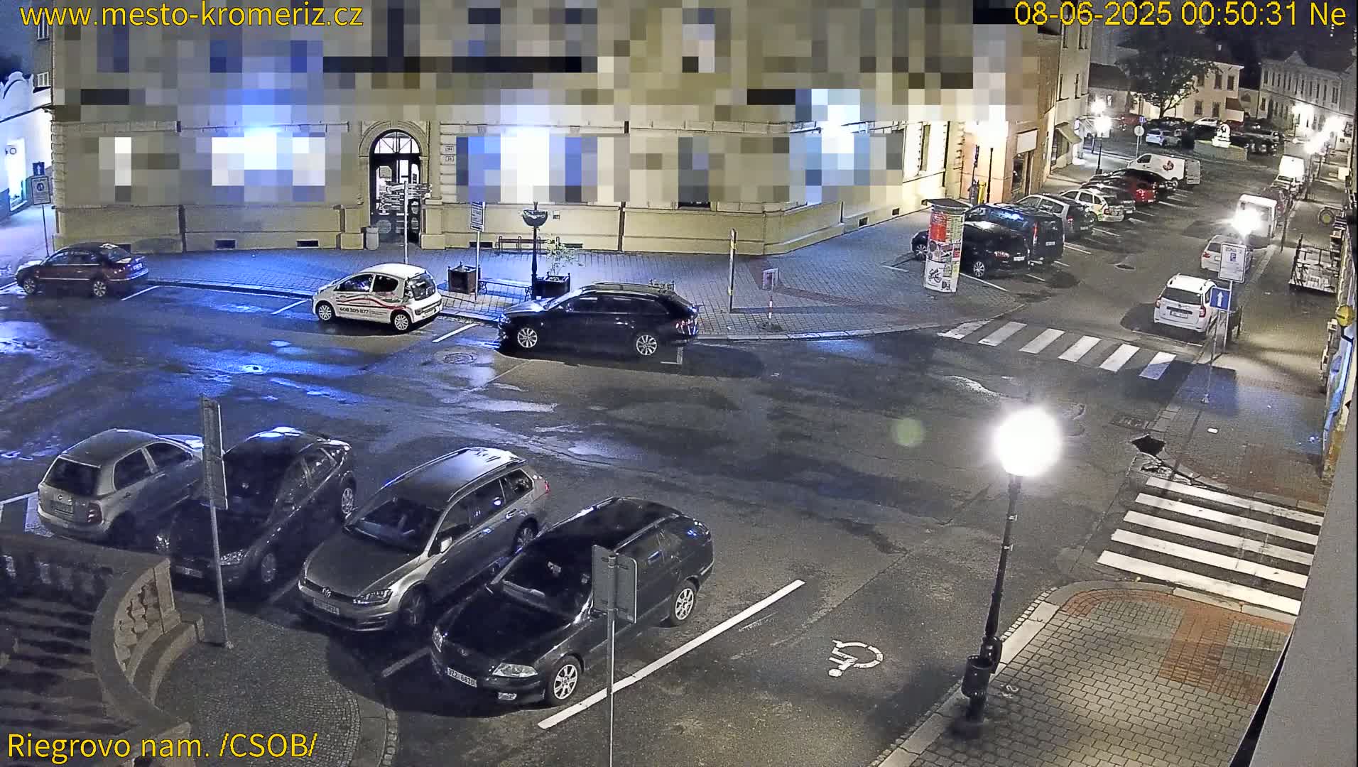 A nighttime, wet street scene shows several cars parked in a lot next to a building, with a crosswalk and a lamppost visible.