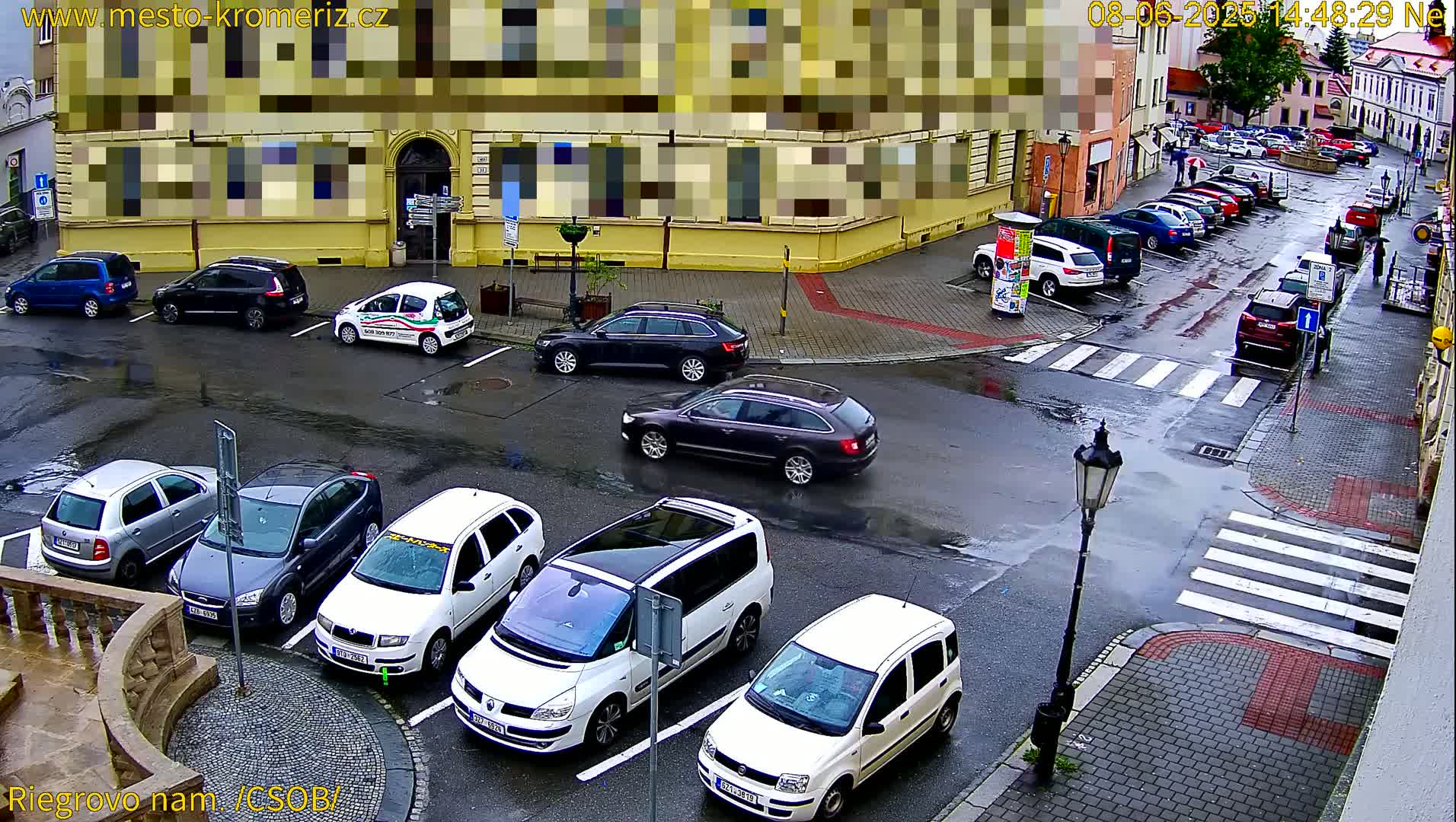 A wet city square with parked cars and a few pedestrians, under an overcast sky.