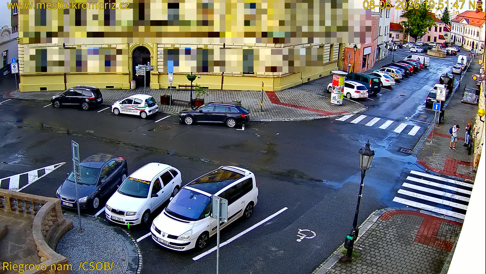A wet city square with several cars parked in a lot, surrounded by buildings under a cloudy sky.