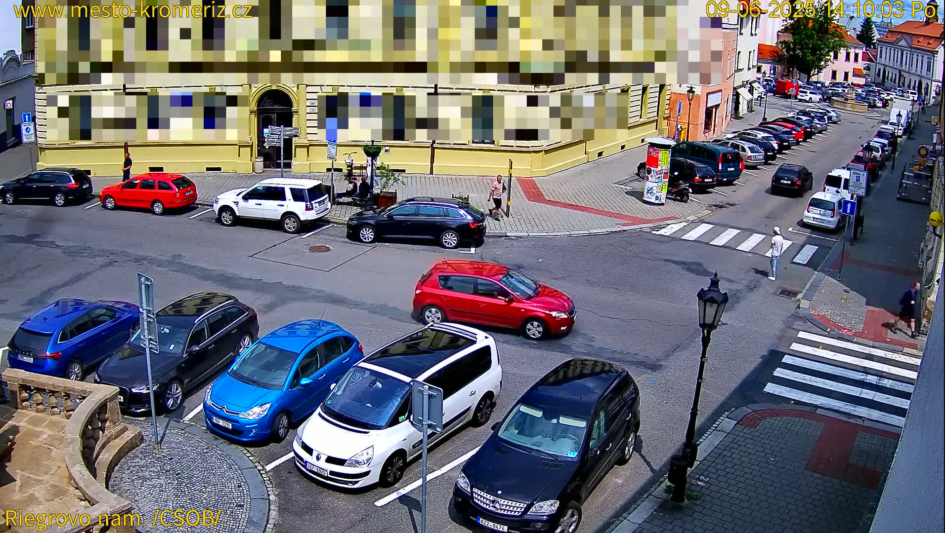 A sunny street scene shows several cars parked in a lot and along the curb near a building, with pedestrians walking on a crosswalk.