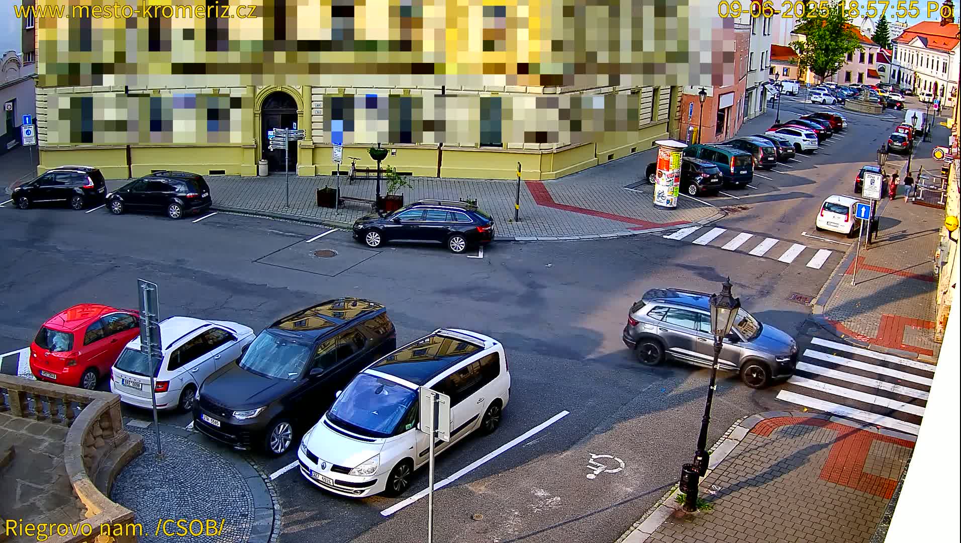 A street corner with parked cars and a few cars in motion, on a sunny day.