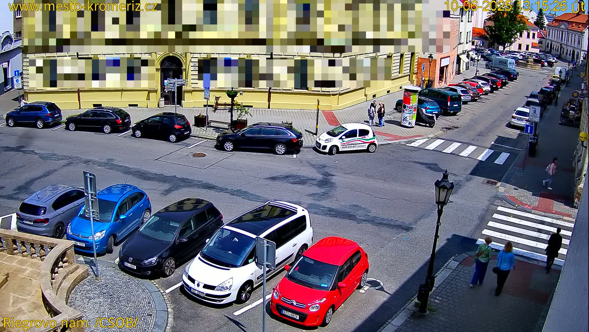 A sunny street scene shows several cars parked in a lot and along a street, with a few pedestrians walking nearby.