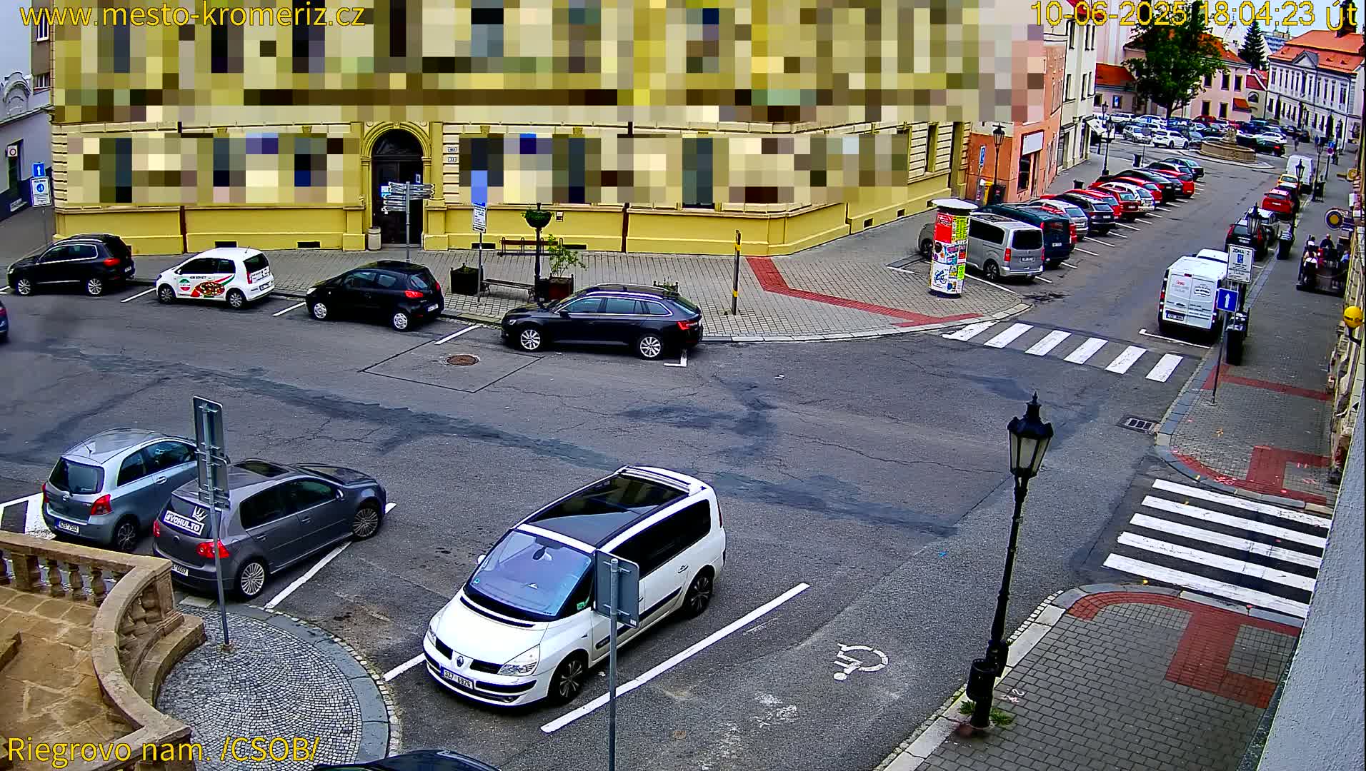 A street corner with parked cars, a white minivan, and a yellow building is shown on a sunny day.