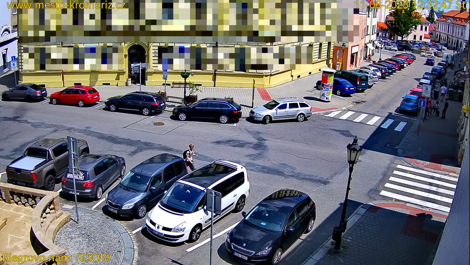 A sunny street scene shows several cars parked in a lot and along the side of a building, with a crosswalk and pedestrians visible.