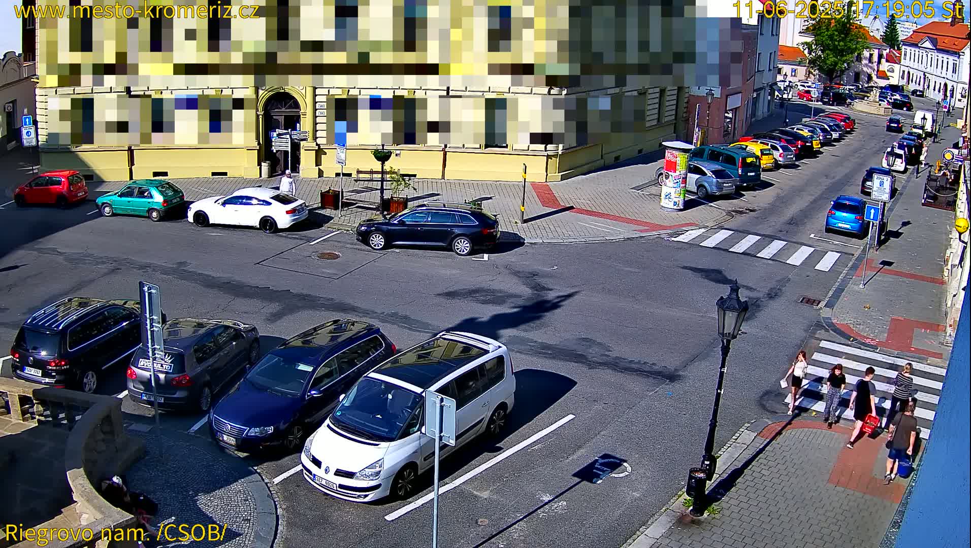 A sunny city street scene shows several cars parked and driving around a corner, with pedestrians crossing a crosswalk.