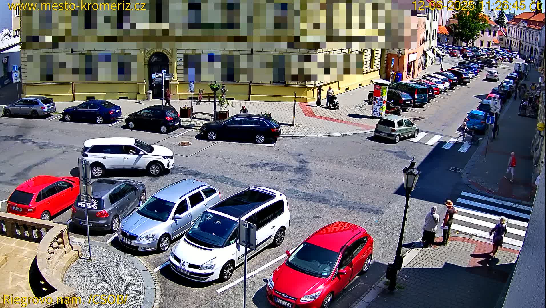 A sunny street scene shows several cars parked along the curb and in a parking lot near a building, with pedestrians crossing a crosswalk.