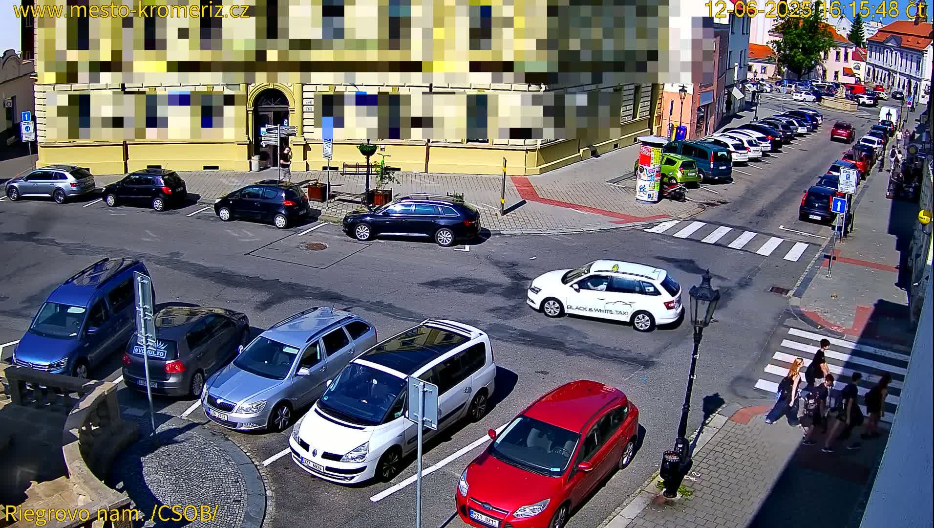 A sunny day in a town square with several cars parked in a parking lot and more cars parked along the street, and a few pedestrians crossing a crosswalk.