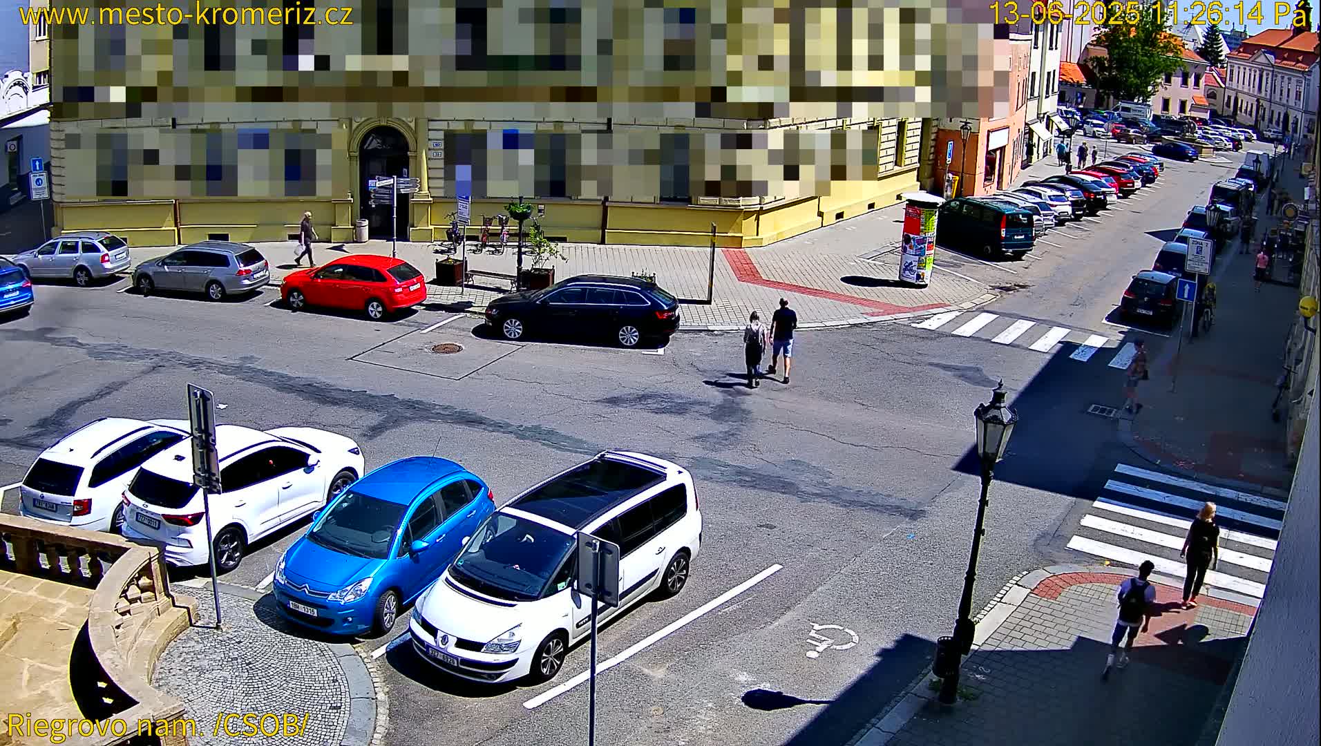 A sunny street scene shows several parked cars and a few pedestrians walking near a crosswalk, with buildings lining the street.