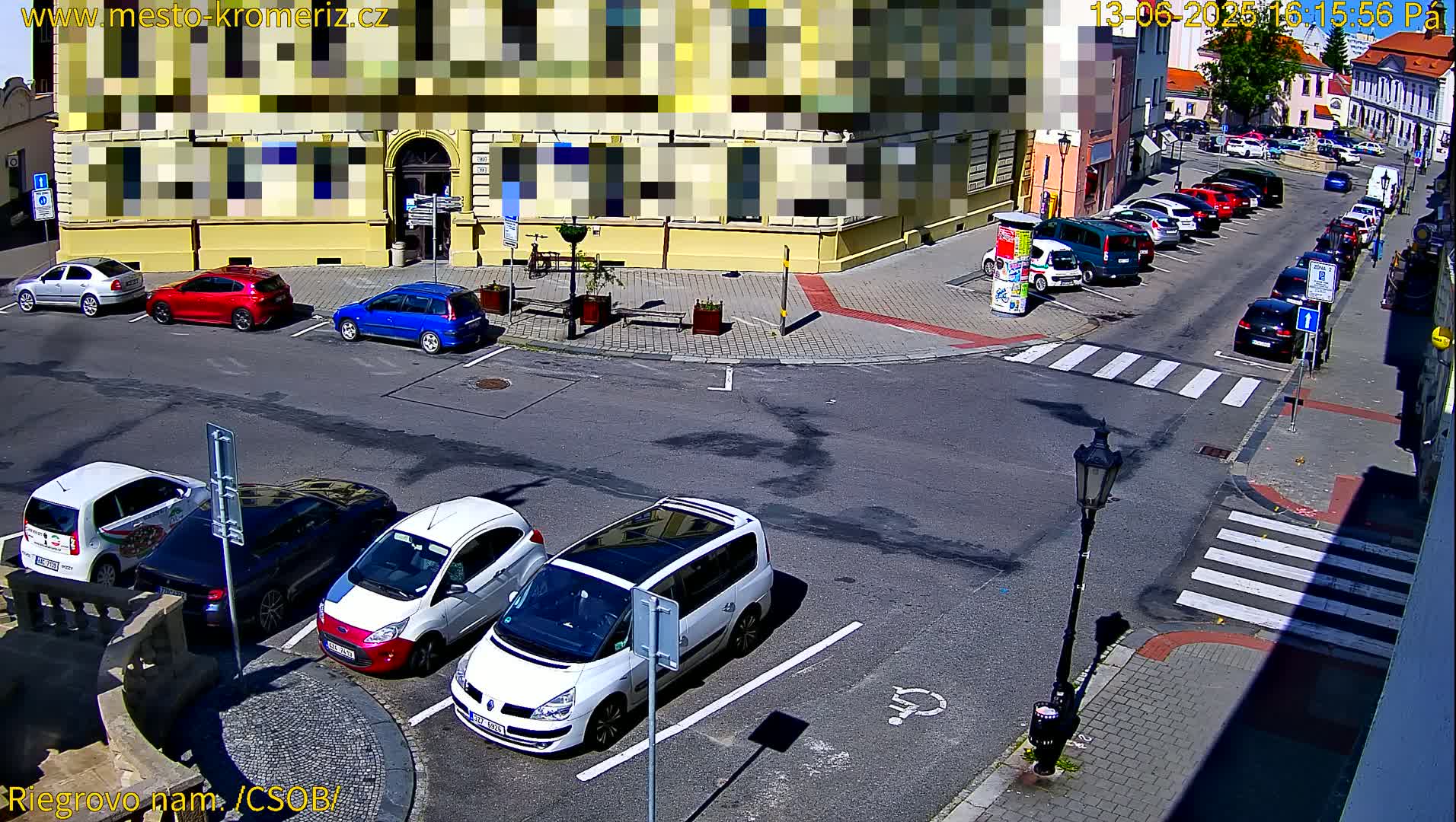 Several cars are parked in a sunny town square near a building, with additional cars parked along a street.