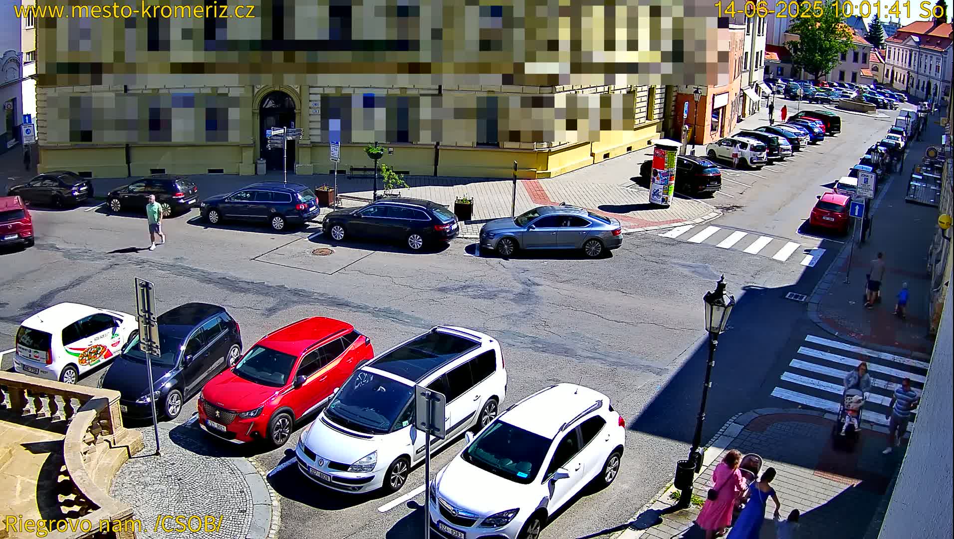 A sunny street scene shows several cars parked in a town square, with pedestrians walking on the sidewalks and crossing a crosswalk.