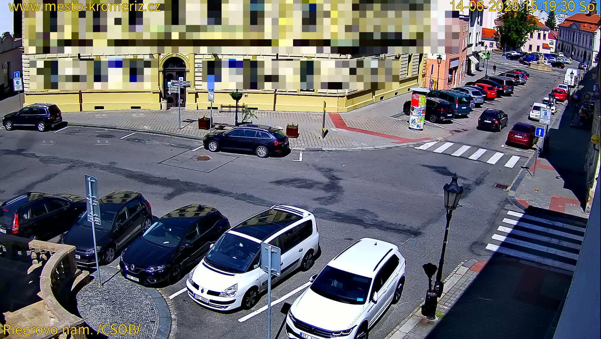 A sunny street scene shows several cars parked in a lot and along the curb, near a yellow building.