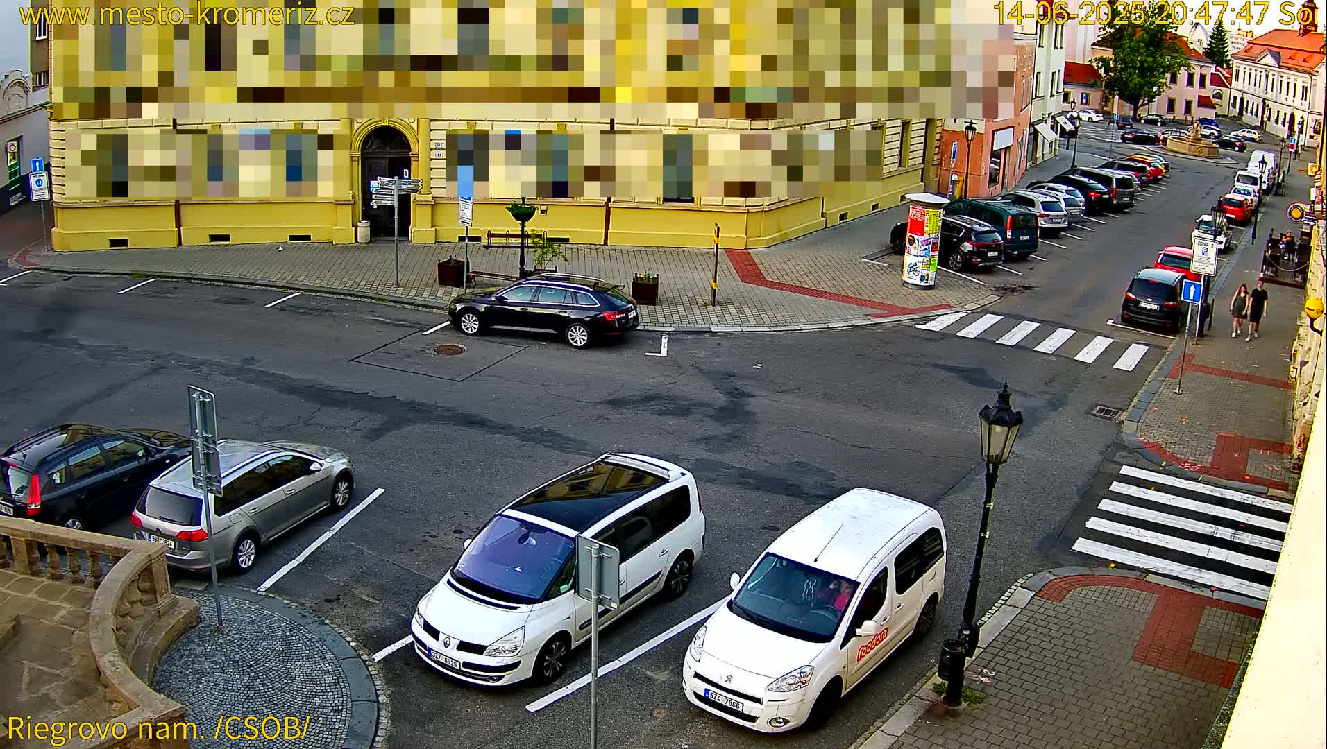 A sunny street scene shows several cars parked in a lot and along the curb, near a yellow building.