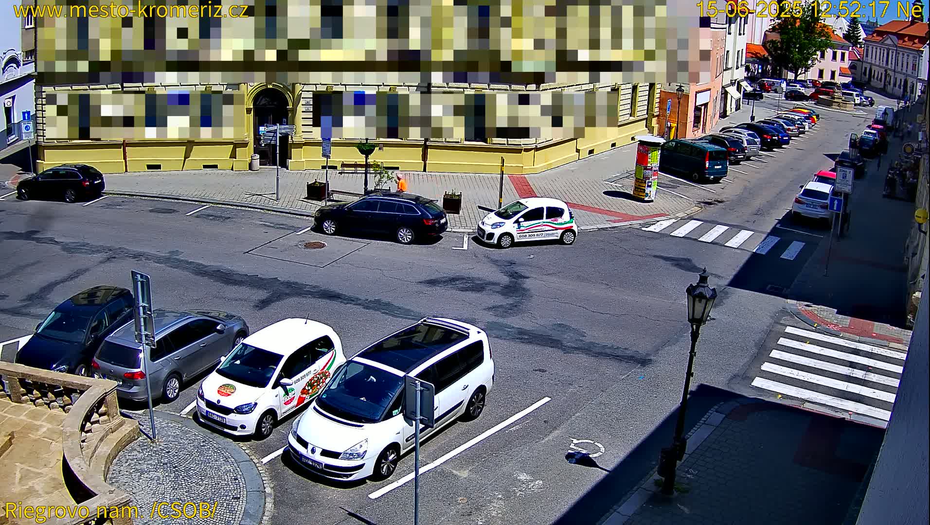 A sunny street scene shows several cars parked in a lot near a building and a crosswalk, with other cars driving down the road.