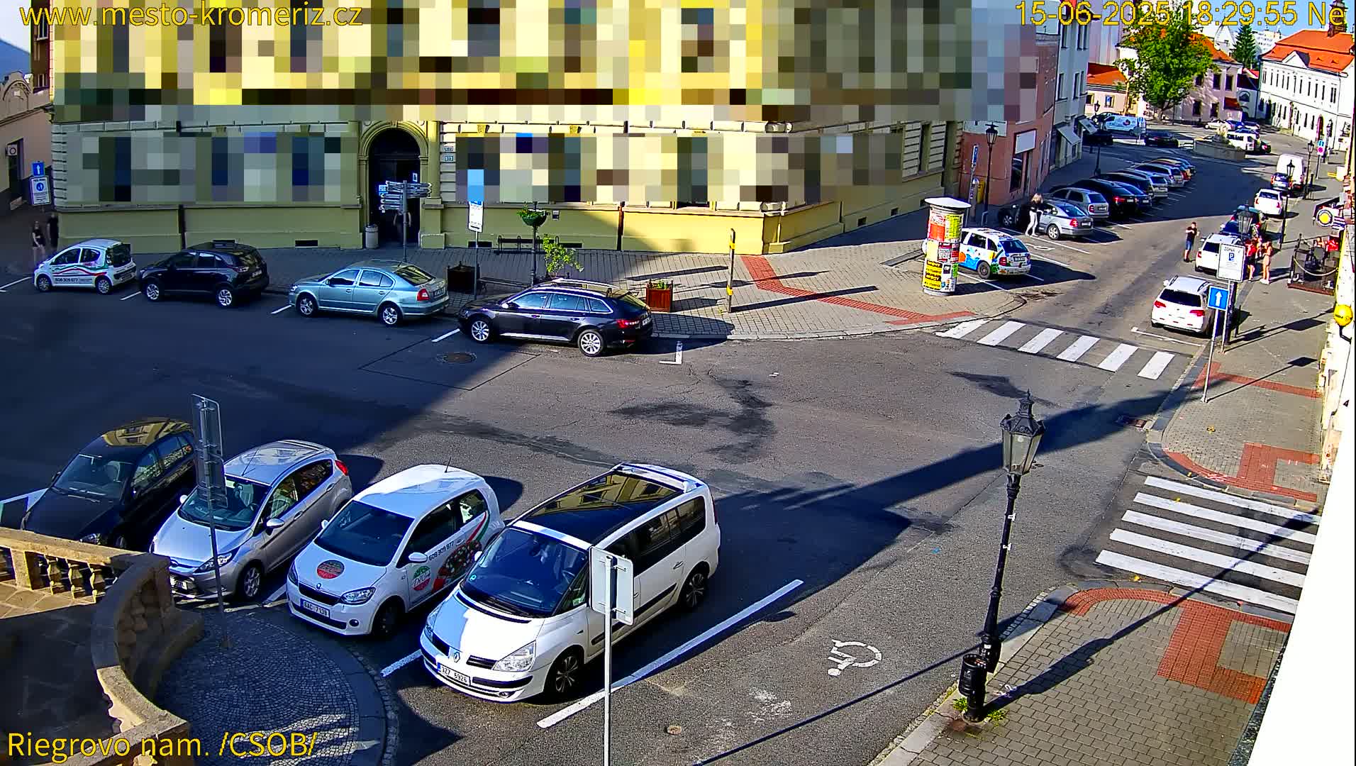 Several cars are parked on a sunny street corner near buildings, with some cars driving past a crosswalk.