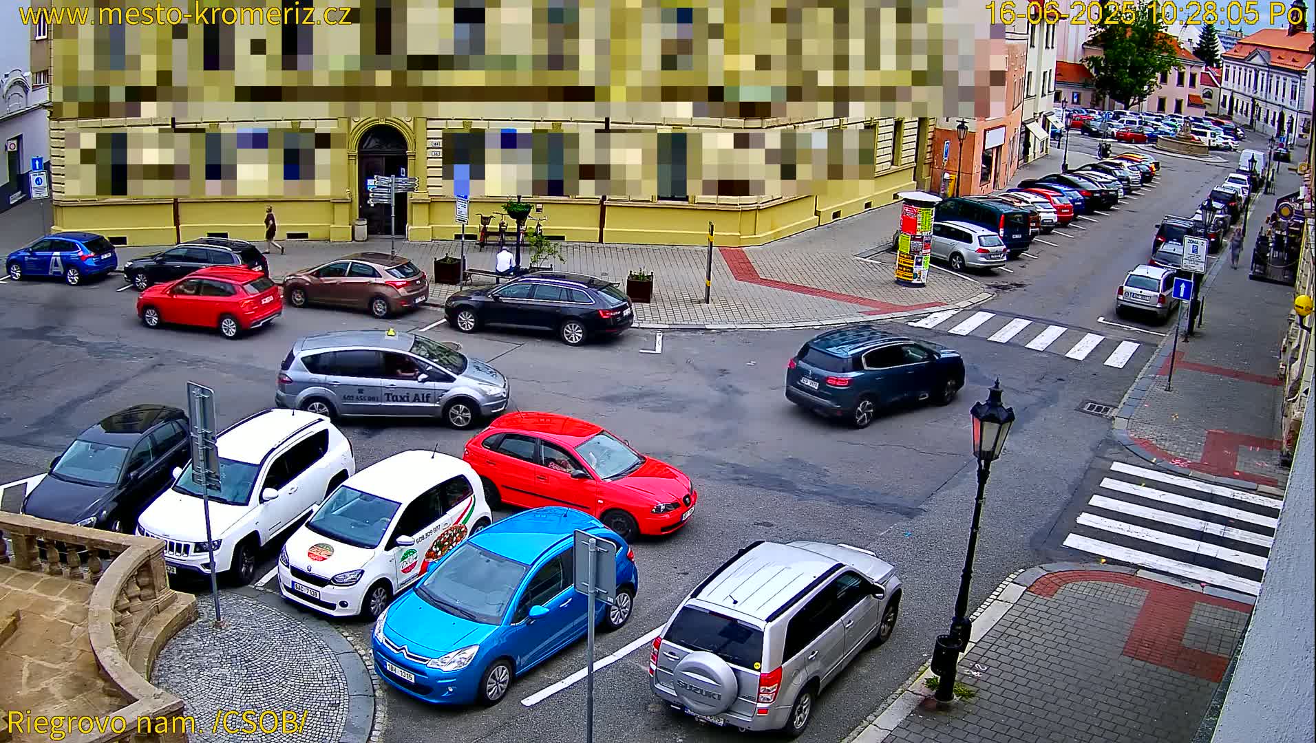 A street intersection with numerous parked cars, some pedestrians, and a taxi, under a partly sunny sky.