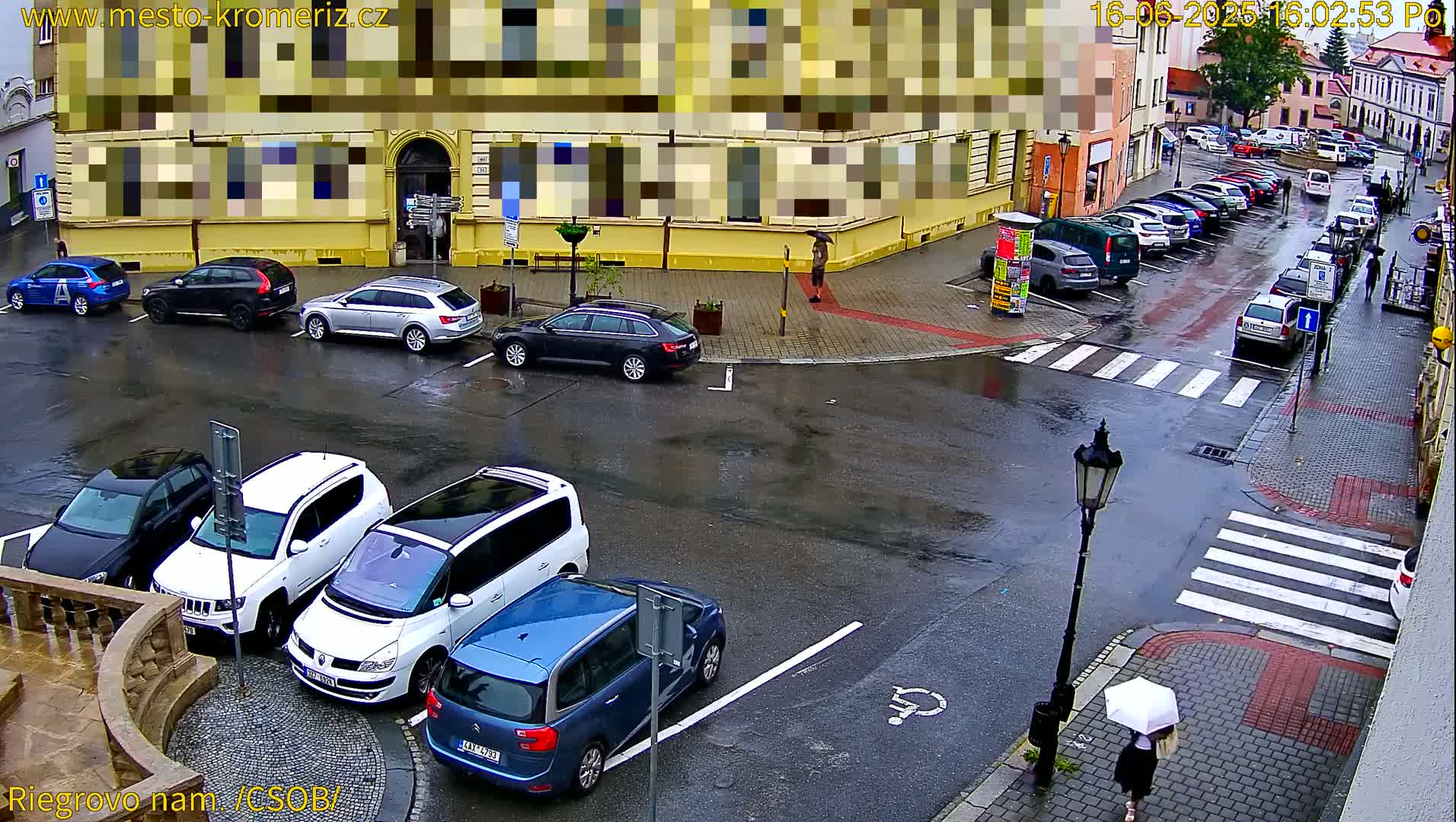 A wet city street scene shows several parked cars around a corner, with a person carrying an umbrella walking on the sidewalk, and a person standing on a corner.